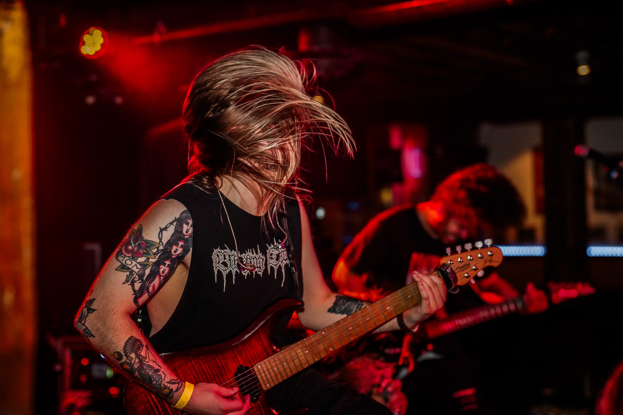 A black and white photo of a live metal band performing on stage with energetic fans in the foreground. The band's setup includes guitars and drums, with bright stage lights illuminating them.