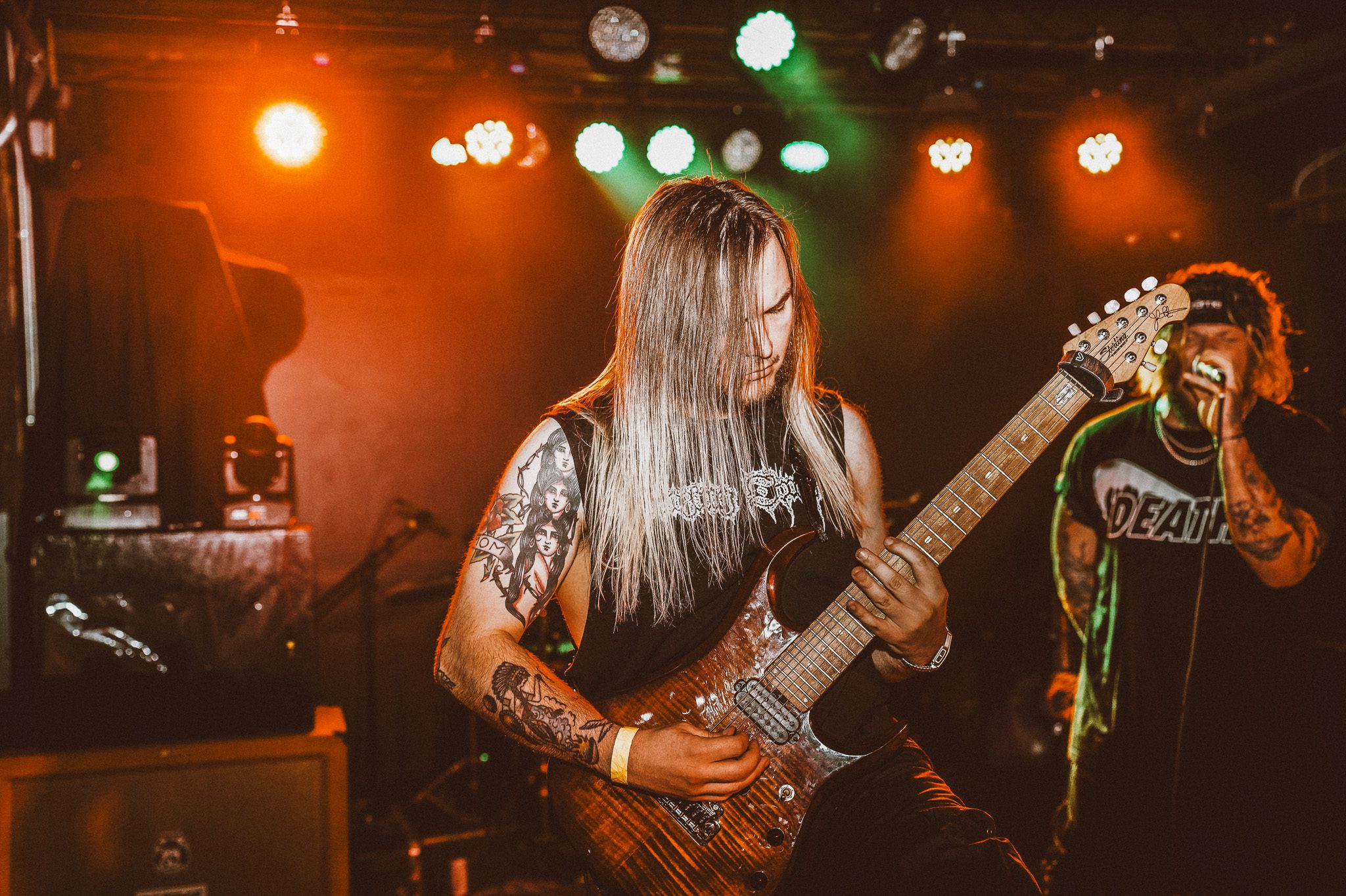 A band performing on stage with green lights, featuring a vocalist with a microphone, a drummer, and two guitarists. The atmosphere is energetic and lively, typical of a live metal performance.