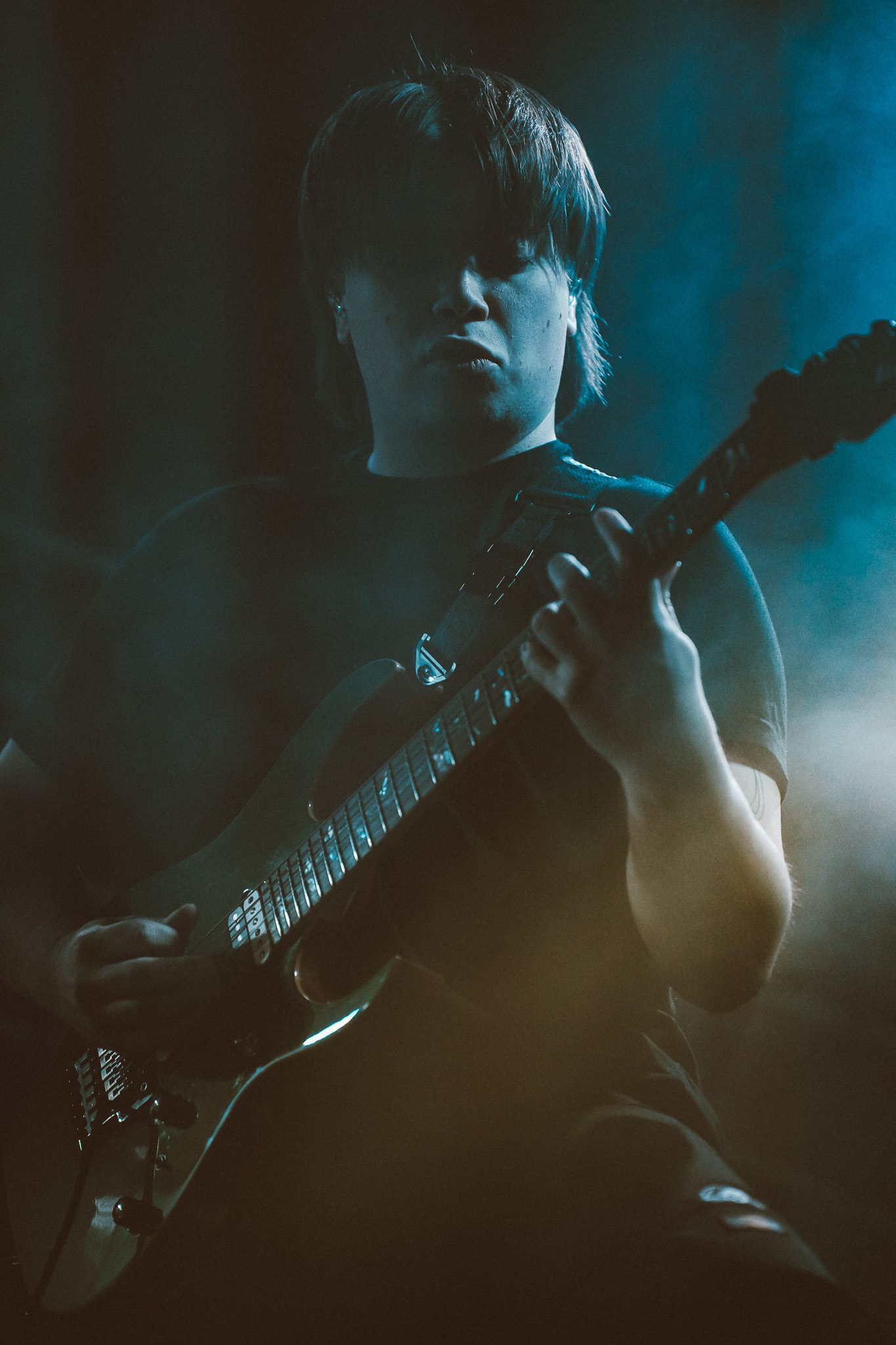 A musician playing a light blue bass guitar on stage, dressed in a gray hoodie, surrounded by amplifiers in a dimly lit environment.