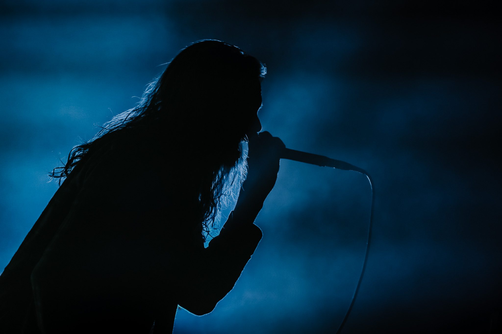 A metal vocalist passionately performing on stage, wearing a plaid shirt and a cap, with dramatic lighting and cables in the background.