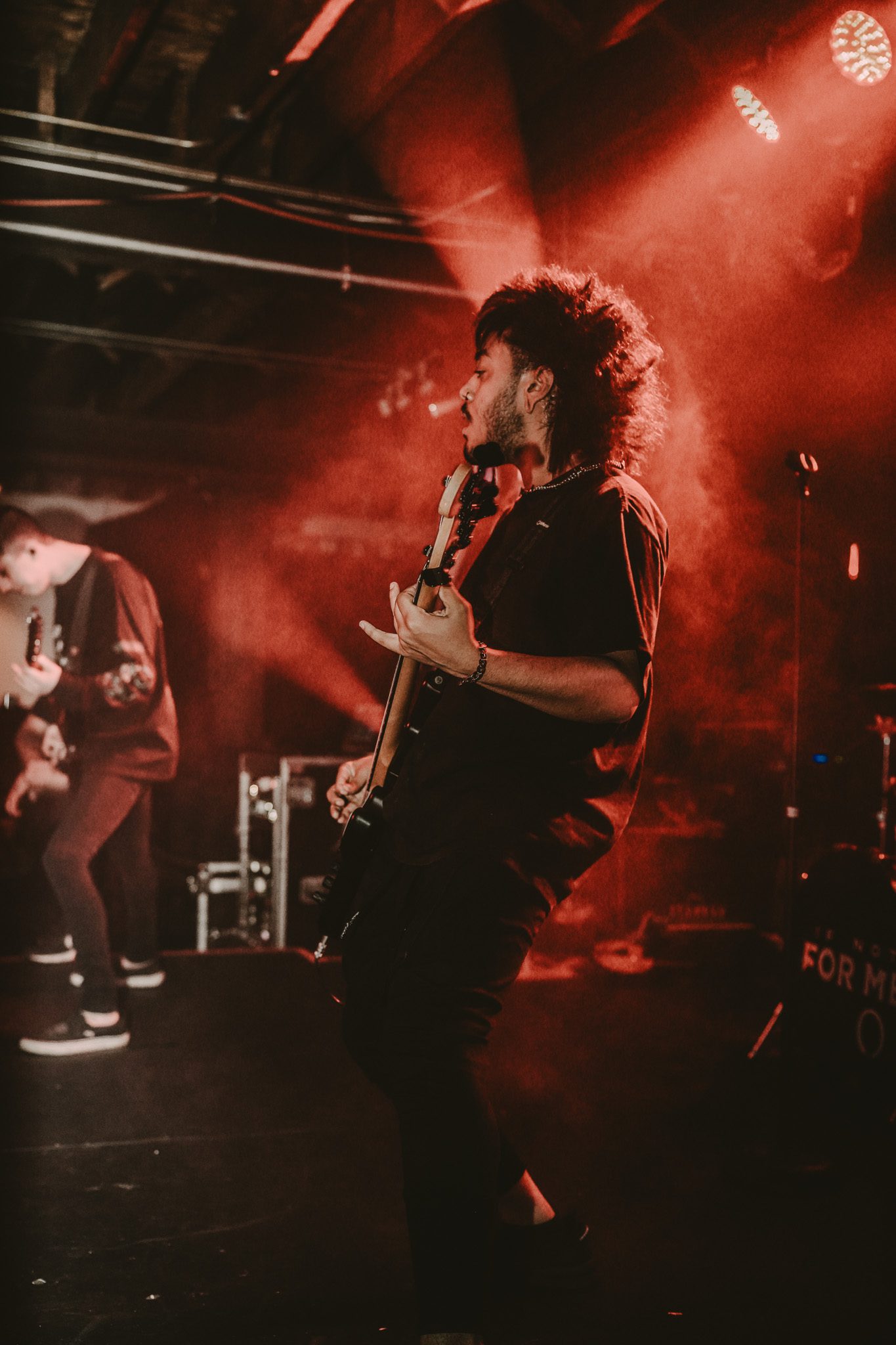 A guitarist performing energetically on stage, with dramatic lighting and a blurred background featuring band equipment.