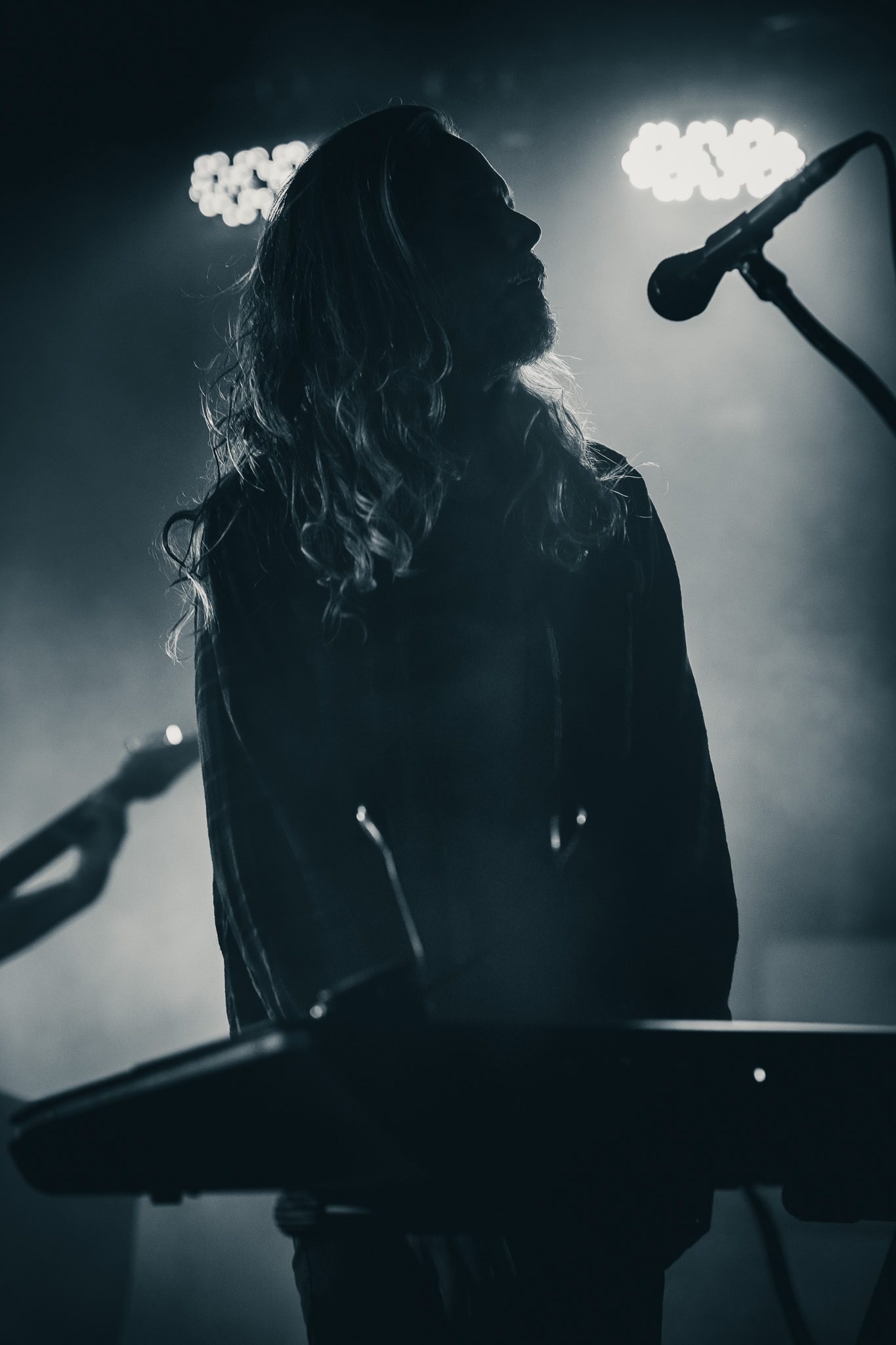 A vocalist passionately performing on stage with a microphone, while a drummer can be seen in the background, surrounded by stage lights and fog.