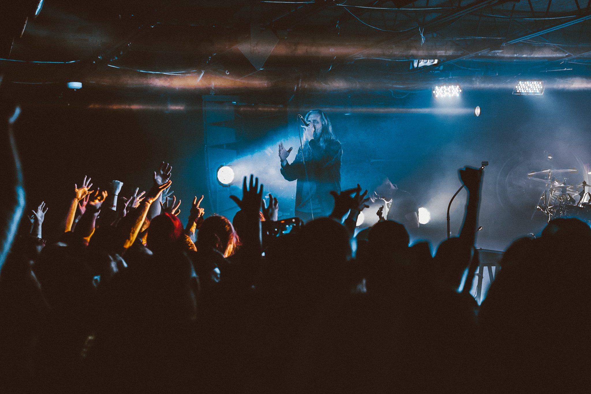 A guitarist performing on stage, wearing a jacket with a lightning pattern, focused on playing an electric guitar, with a drummer visible in the background.