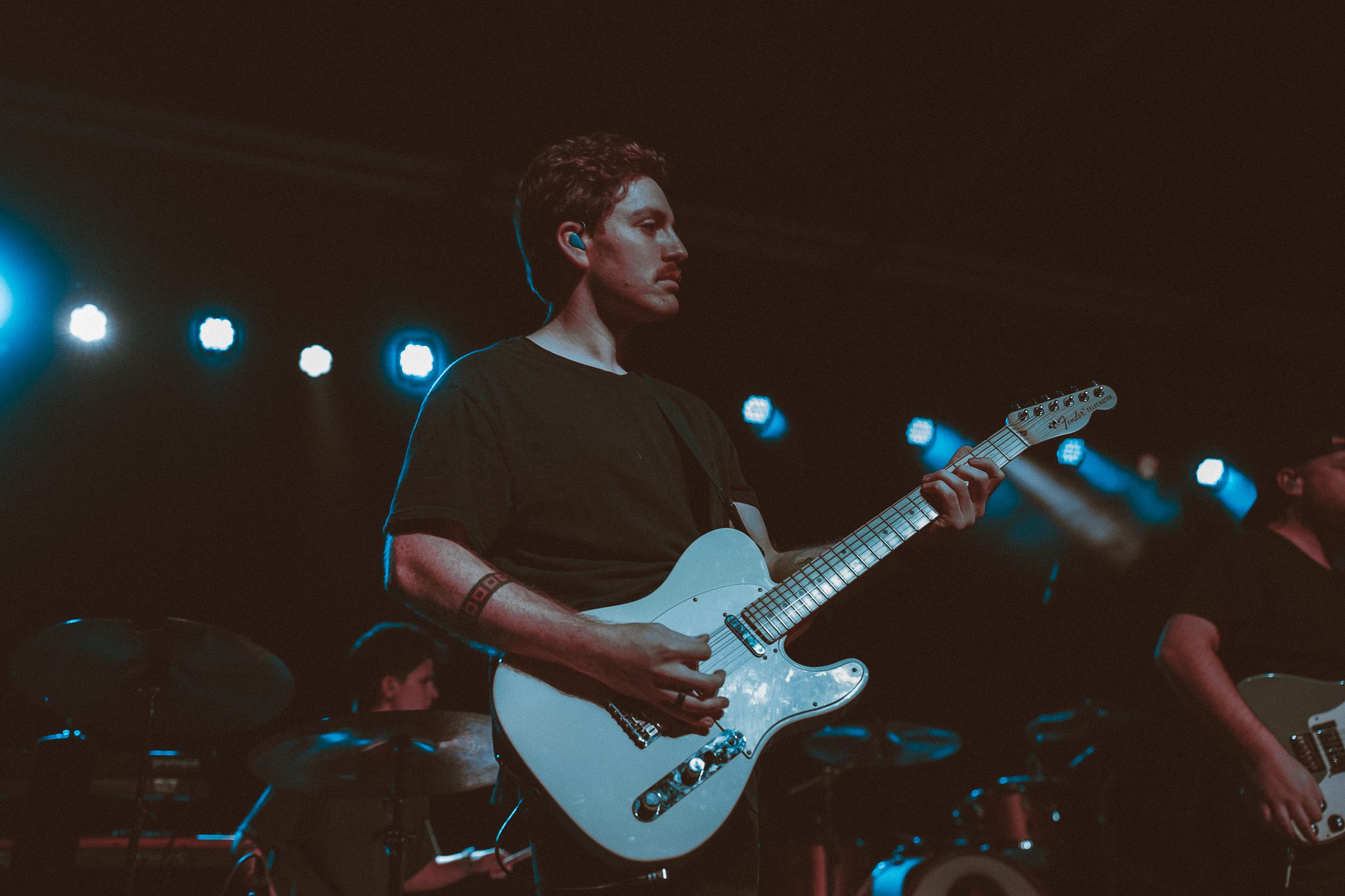 A musician performing on stage with a guitar, set against a red-lit backdrop.