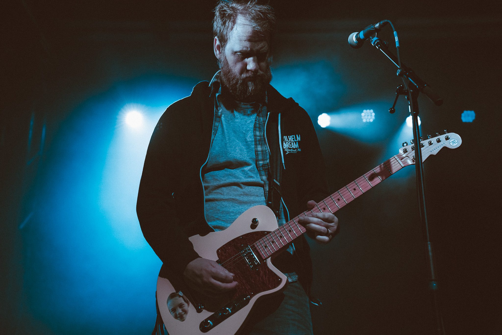 A musician playing an electric guitar on stage at a live concert, with colorful lighting and visual displays in the background.