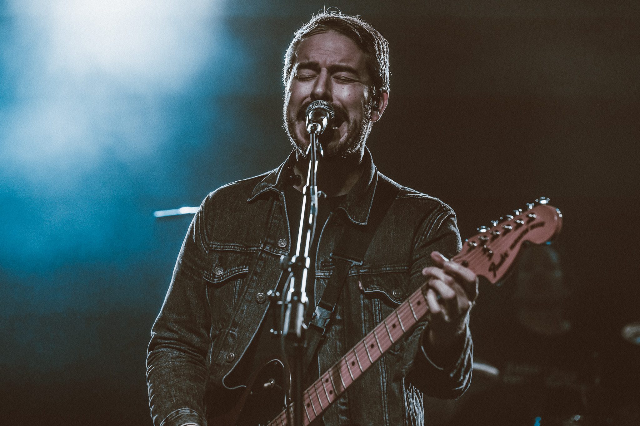 A musician playing the bass guitar on stage, wearing a black t-shirt and a green beanie, with colorful stage lights in the background.