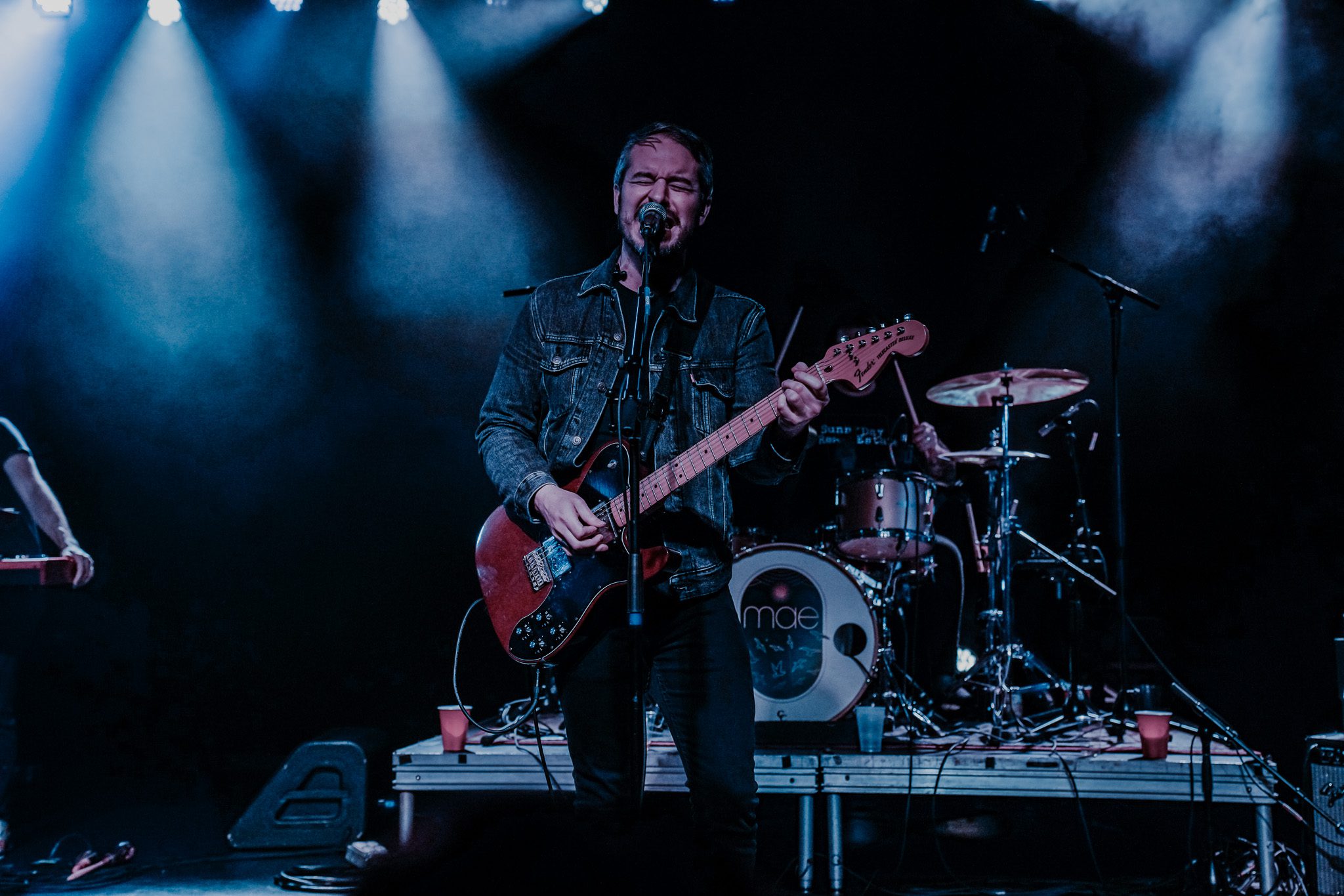 A musician playing a bass guitar on stage, partially obscured by smoke and colored lights.