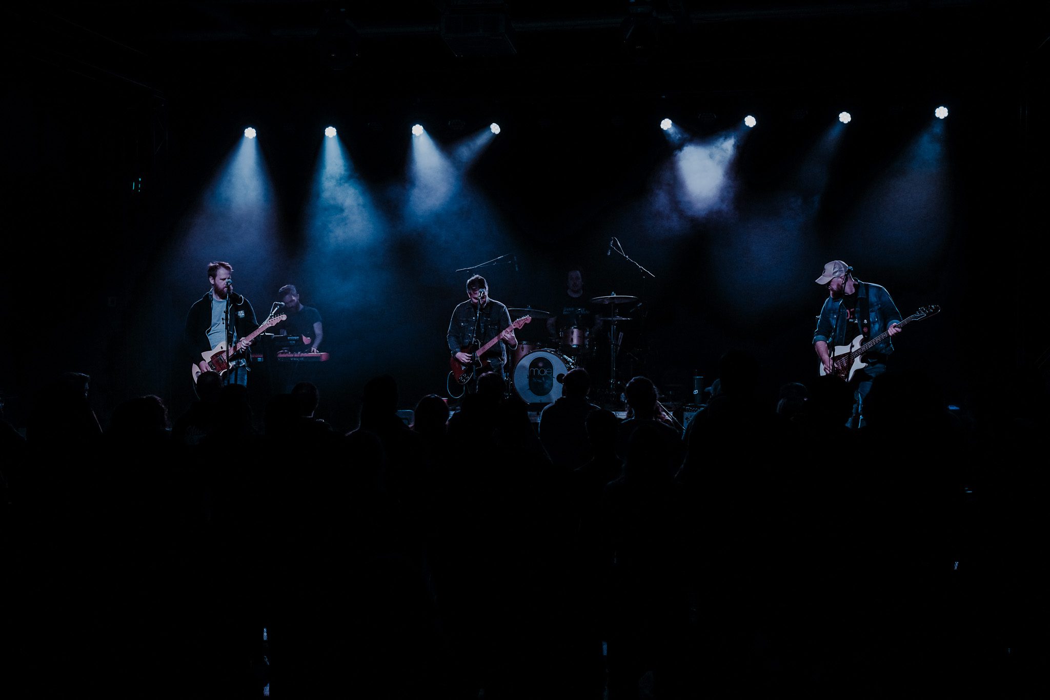 A musician performs energetically on stage, playing an electric guitar with a colorful backdrop and a drummer visible in the background.