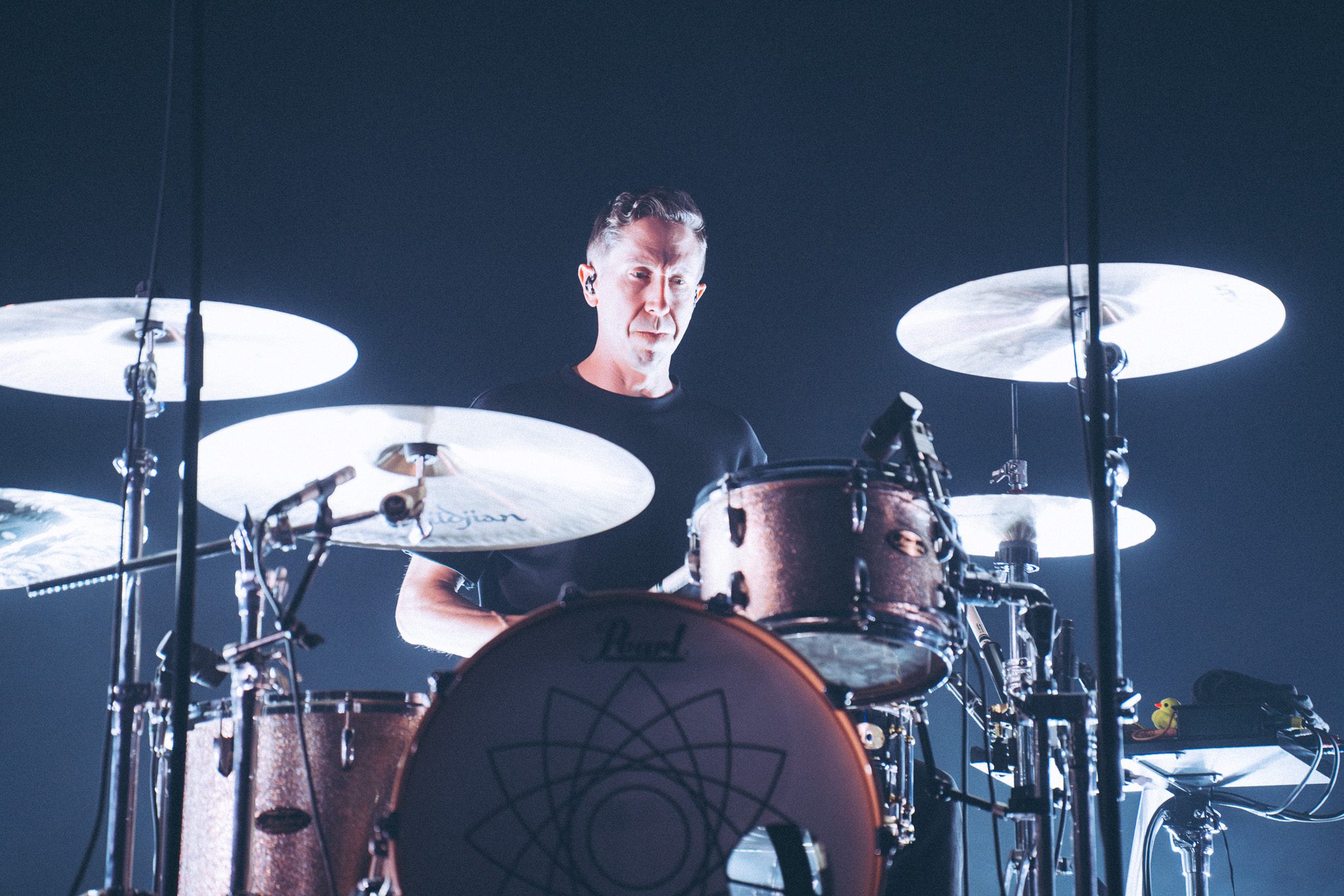 A drummer performing on stage with a focus on the drum set, illuminated by stage lighting.
