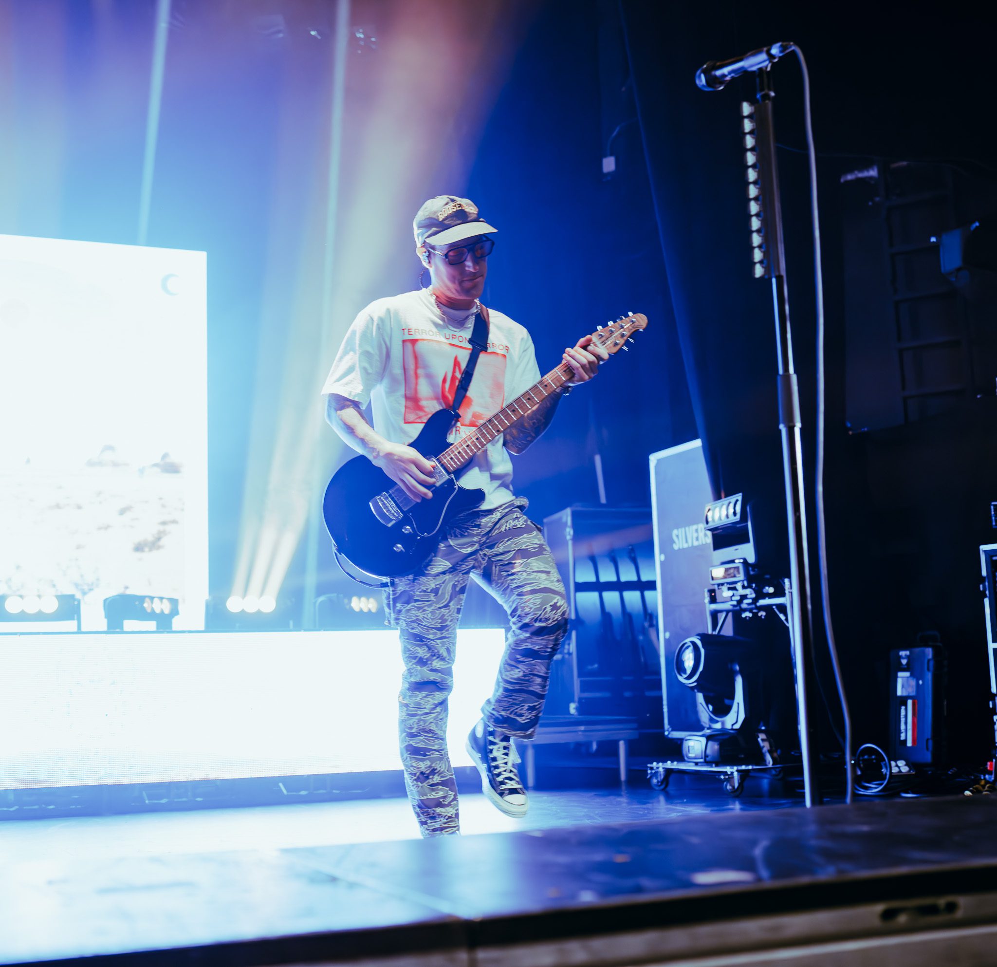 A musician in a white shirt and camo pants plays an electric guitar on stage, with vibrant lighting and a backdrop display.