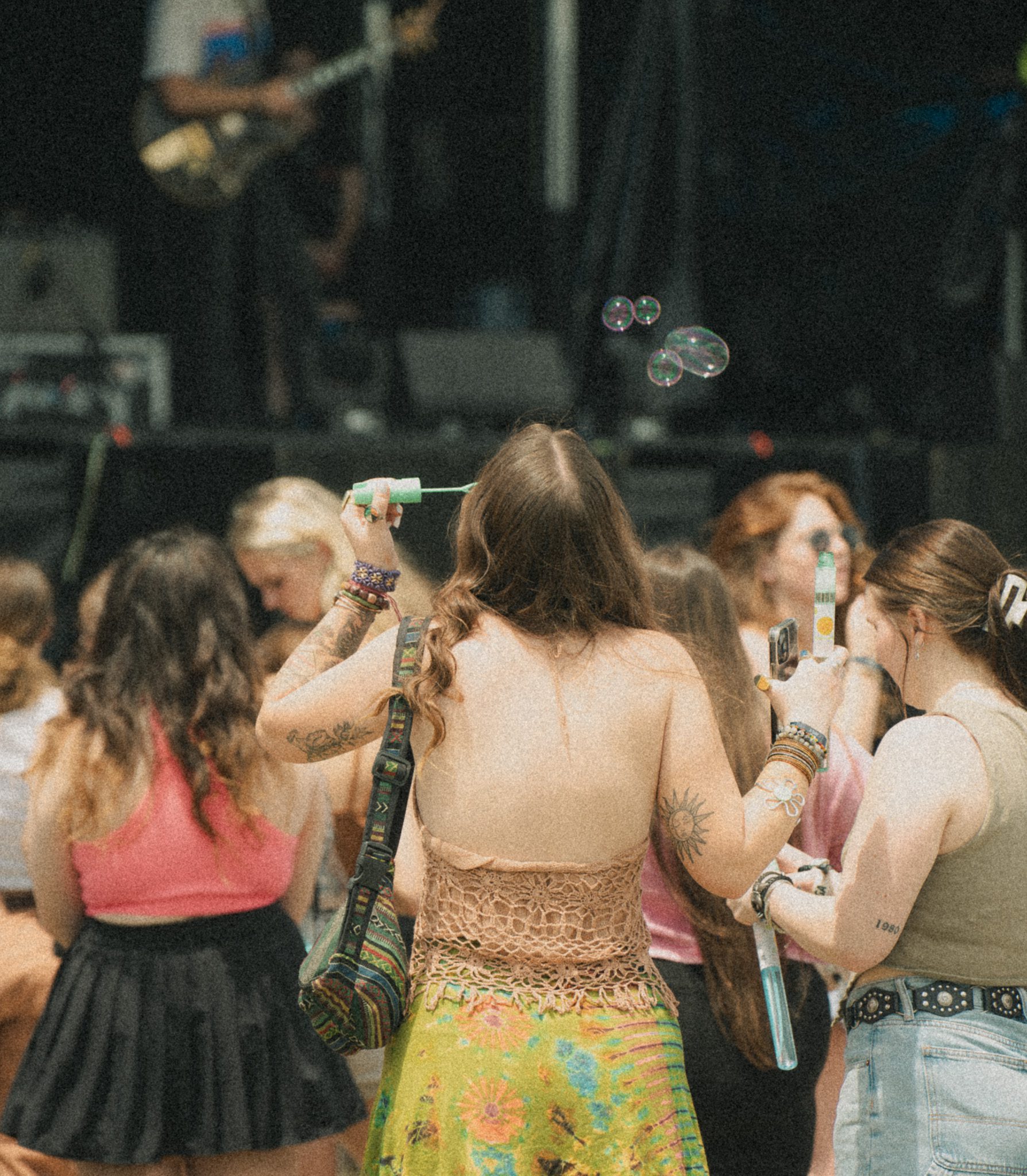 A woman with long hair and a colorful skirt blowing bubbles at a festival, with festival-goers visible in the background enjoying the atmosphere.