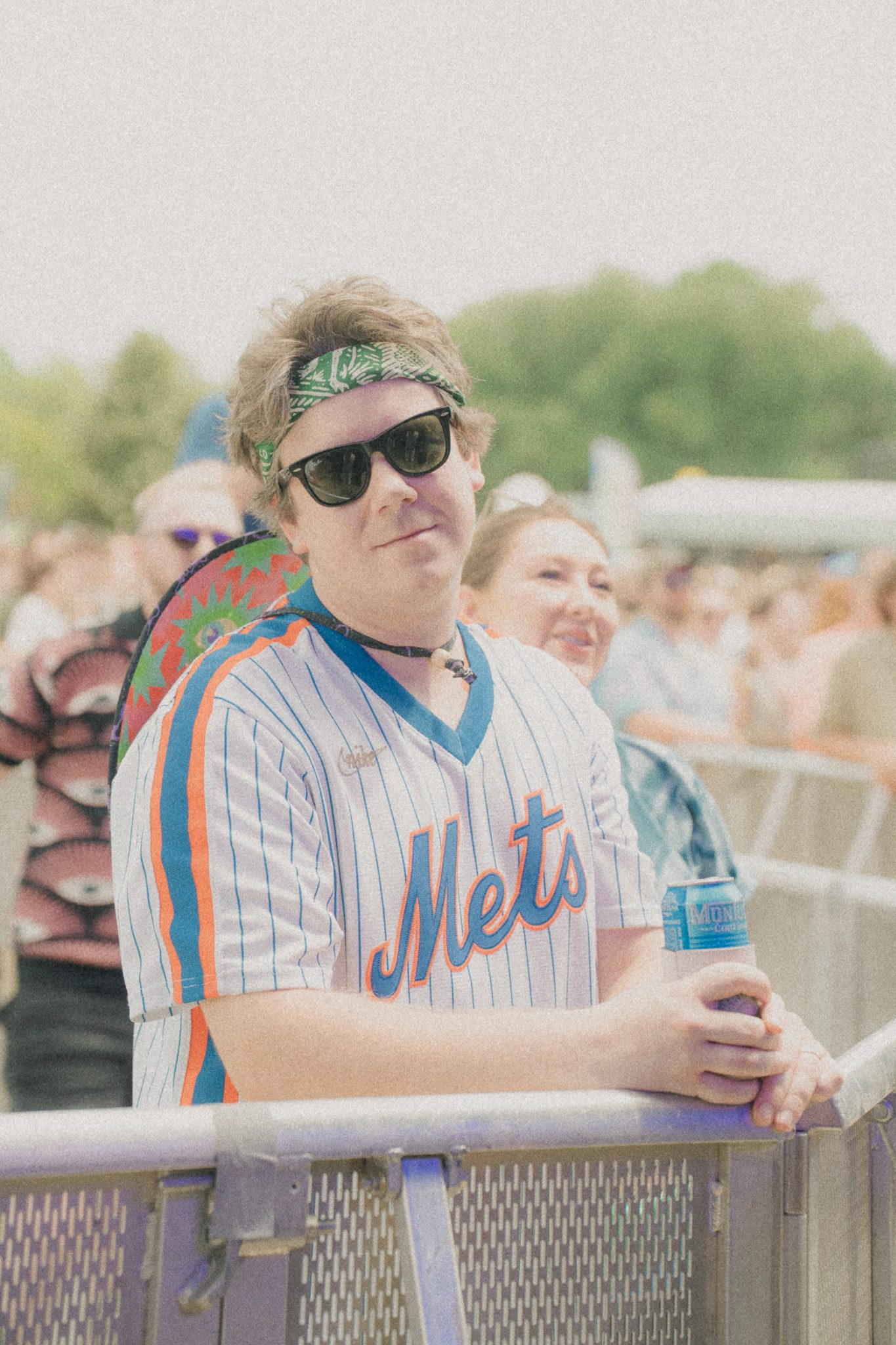 A festival attendee wearing sunglasses and a Mets jersey, standing by the barrier with a can in hand, while smiling in a crowd.