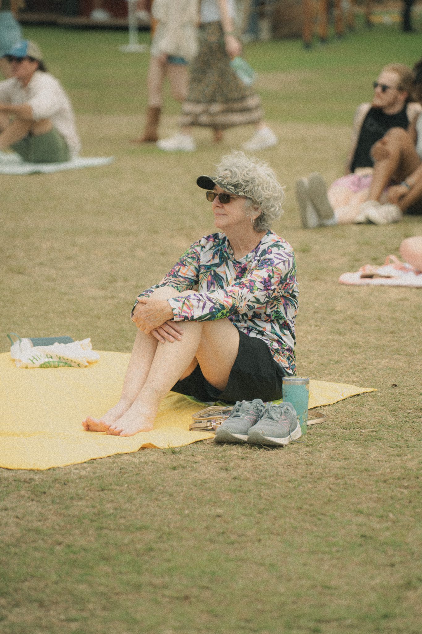 A woman sitting on a yellow blanket at a festival, wearing a colorful shirt and sunglasses, with bare feet and a drink beside her.