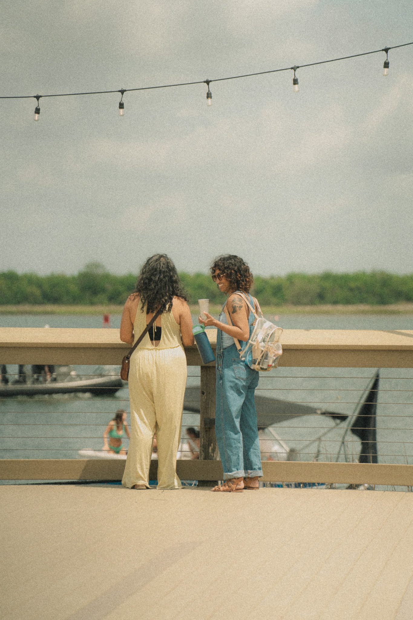 Two women standing on a dock, one wearing a yellow jumpsuit and the other in denim overalls, smiling and enjoying their time together by the water.