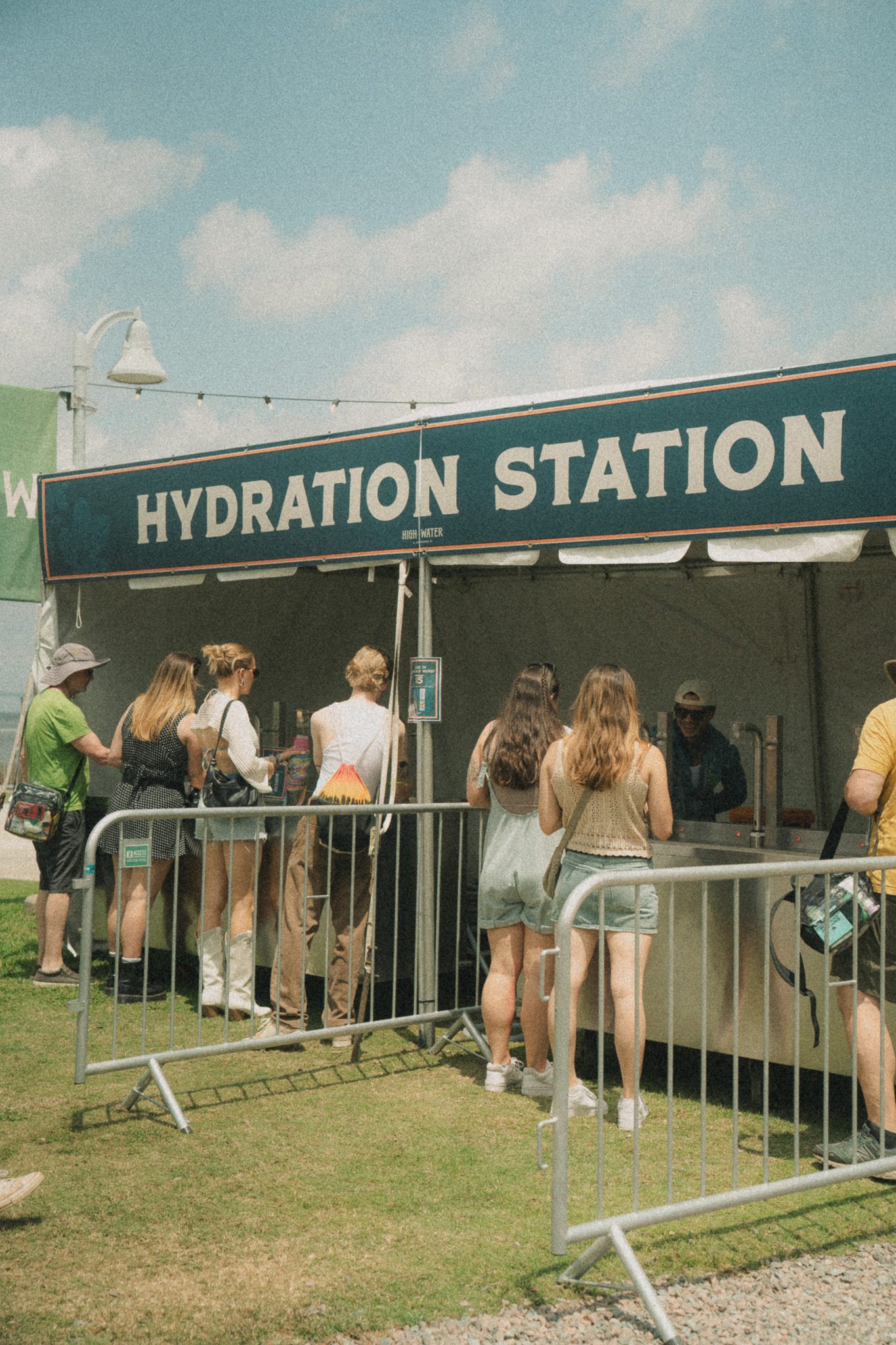 A group of festival attendees lining up at a hydration station under a blue sky, with a sign reading 'HYDRATION STATION' in the background.