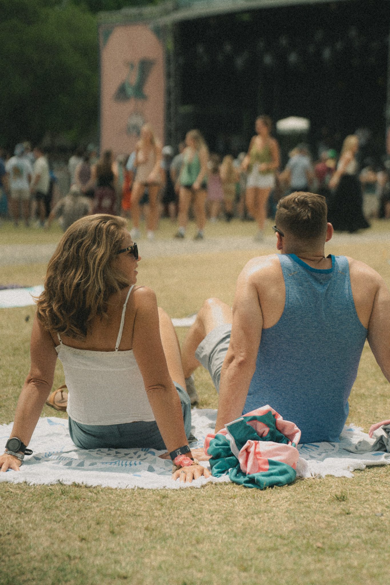 A couple sitting on a blanket at a festival, enjoying the atmosphere with a stage in the background.
