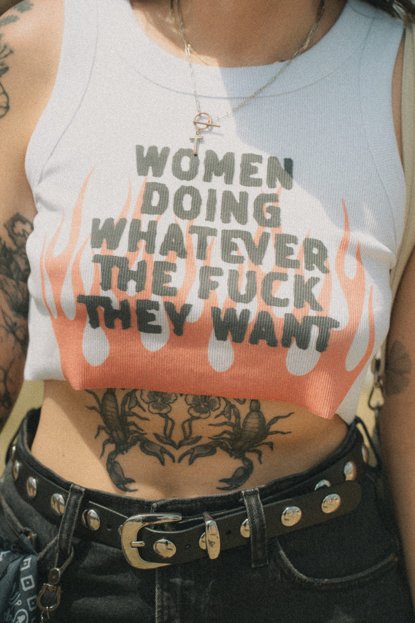 Close-up of a person wearing a sleeveless crop top with the phrase 'Women Doing Whatever The Fuck They Want' printed in bold text, surrounded by flame graphics. The individual also has visible tattoos and is wearing a belt with decorative studs.