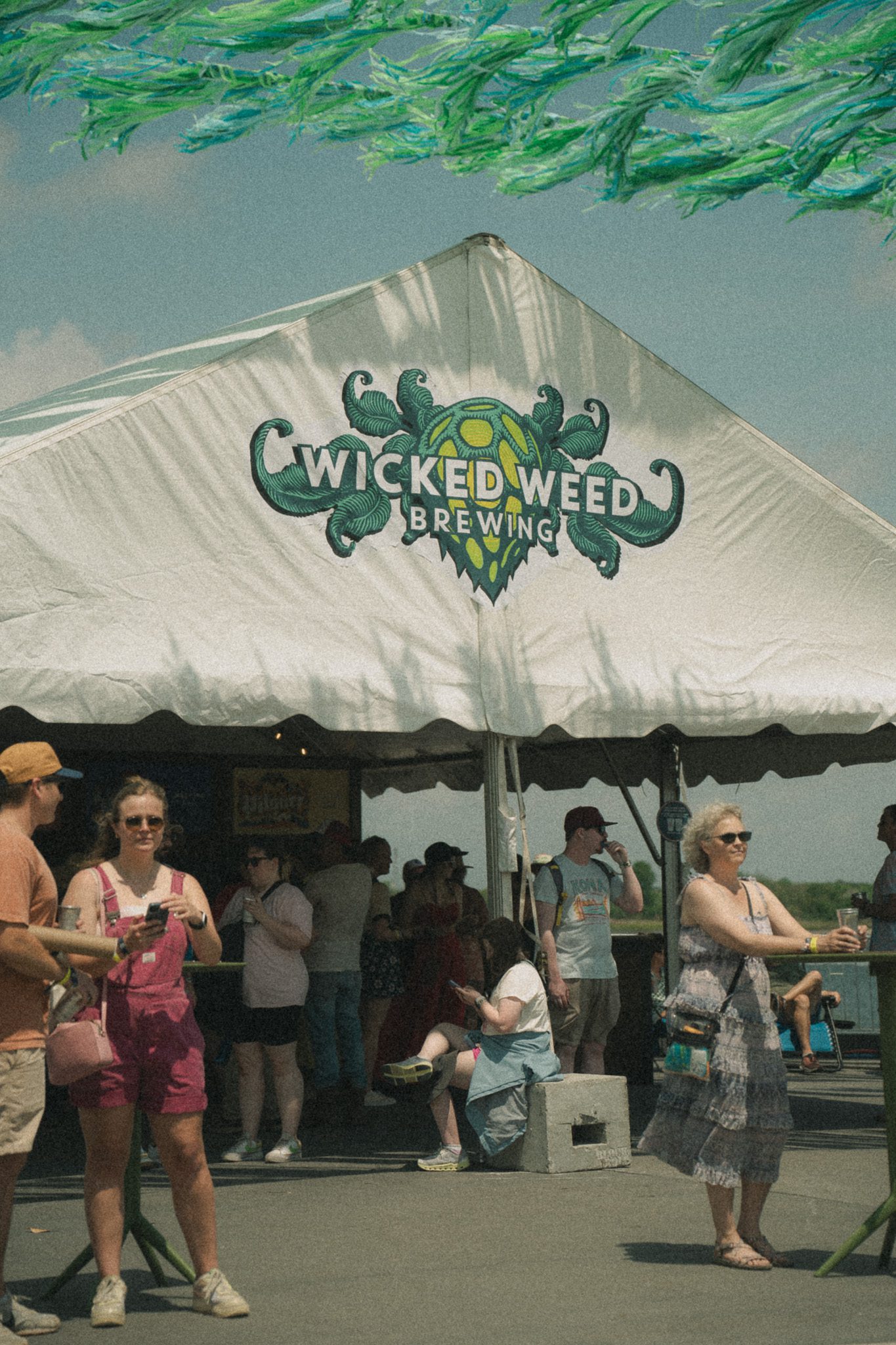 Festival attendees gather near a tent displaying the 'Wicked Weed Brewing' logo, with several people visible in the background enjoying the event.