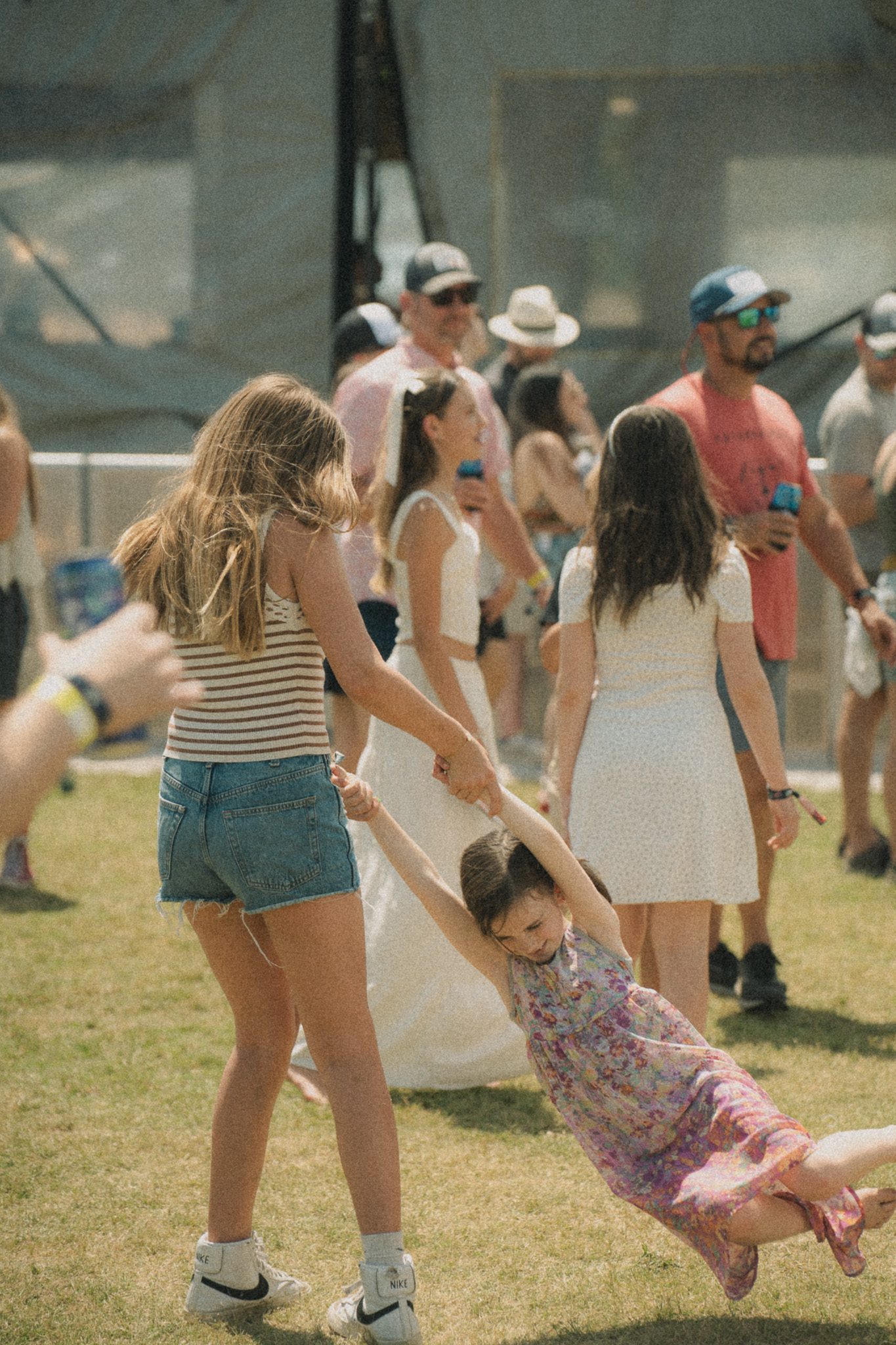 A girl in a striped shirt and denim shorts is joyfully holding hands with a younger girl in a floral dress who is swinging in the air, surrounded by festival-goers in a lively outdoor setting.