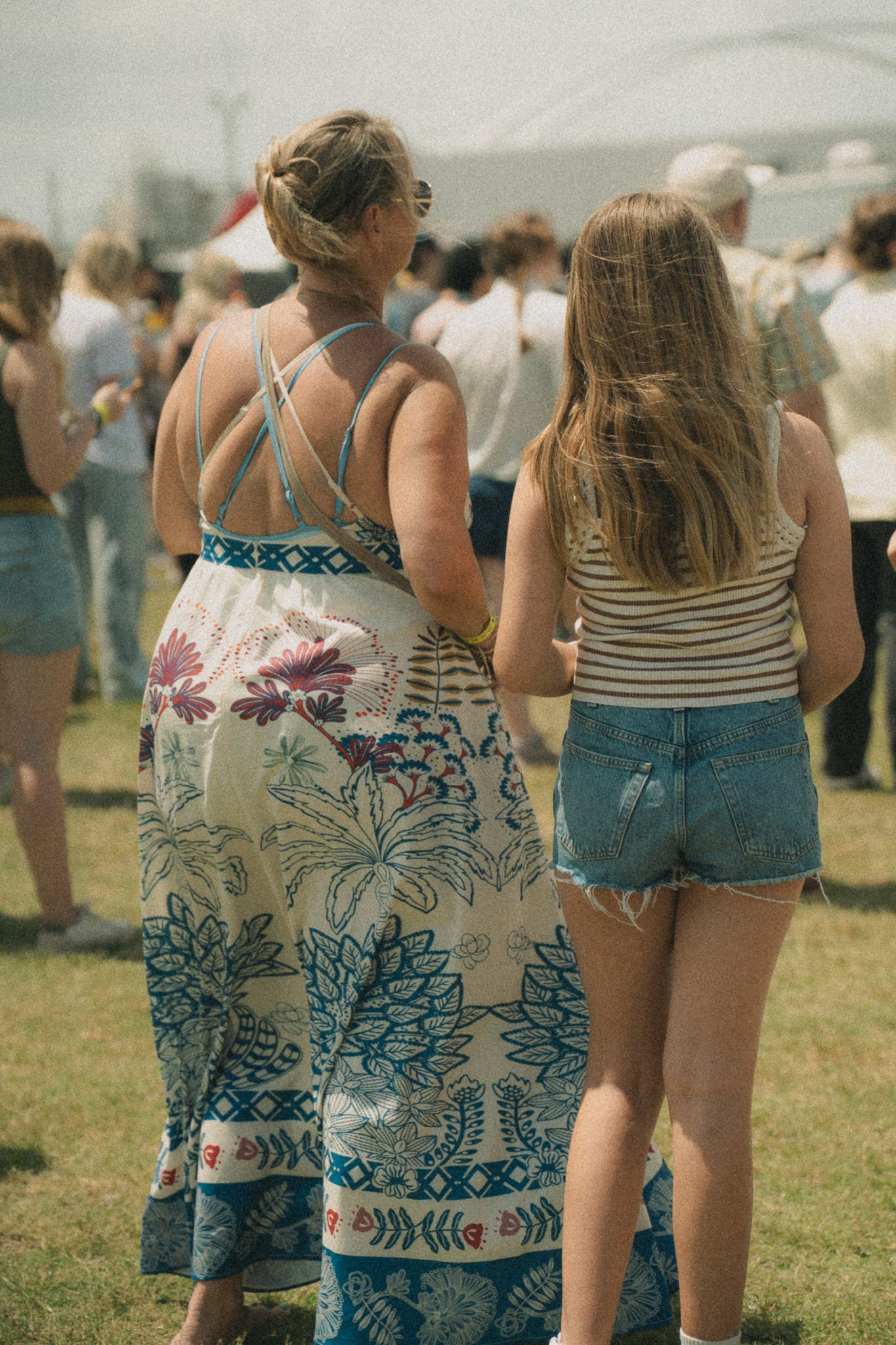 Two women standing side by side at a festival, one wearing a patterned dress and the other in a striped top and denim shorts, enjoying the atmosphere.