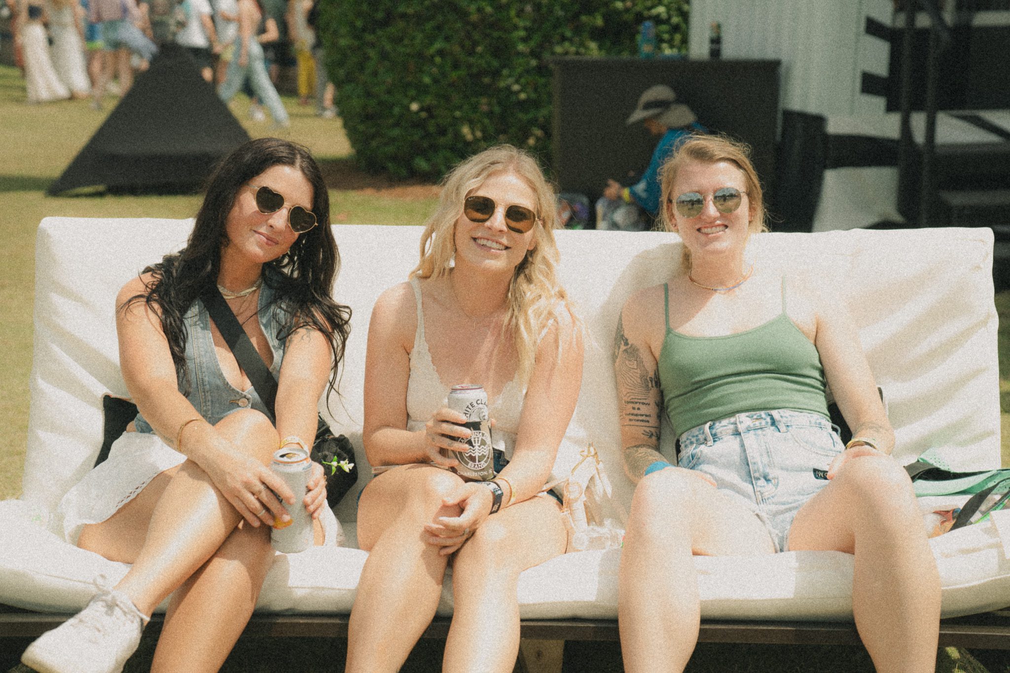 Three women sitting together on a bench at a festival, smiling and holding drinks, with a lively festival atmosphere in the background.