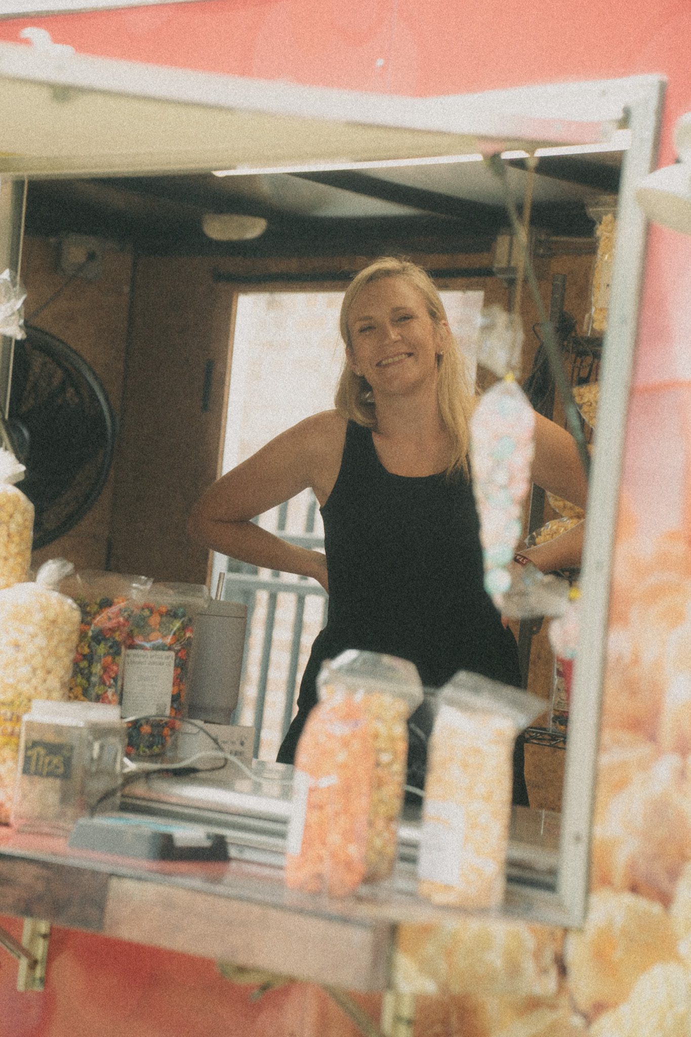 A smiling woman at a festival vendor station, surrounded by colorful candy and popcorn containers.