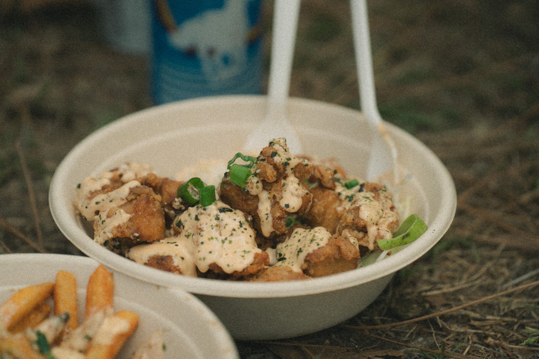 A bowl of flavorful food topped with sauce and green onions, served with two plastic spoons, resting on grass.