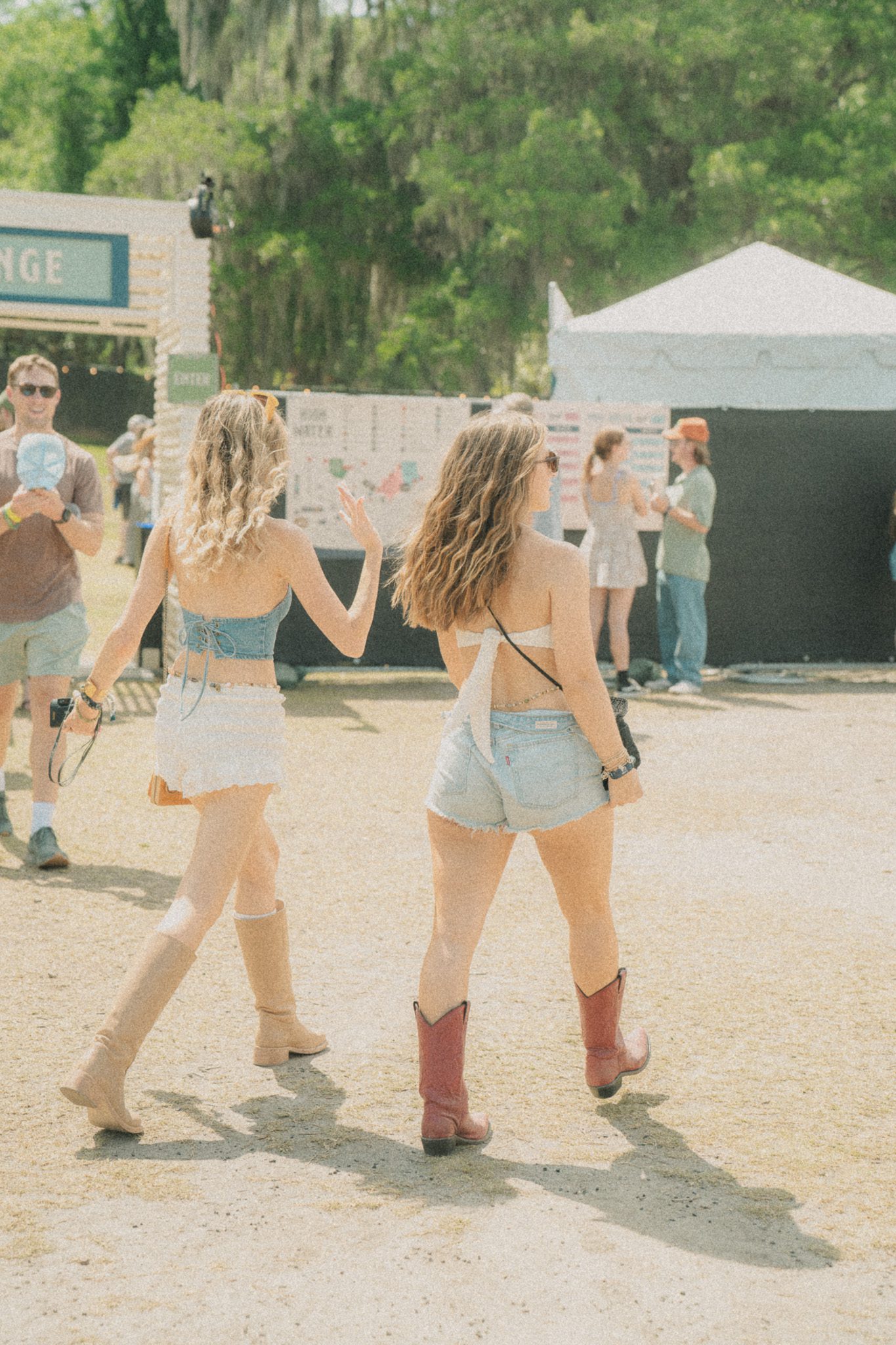 Two women walking away, enjoying the atmosphere of a festival, wearing short denim outfits and cowboy boots.