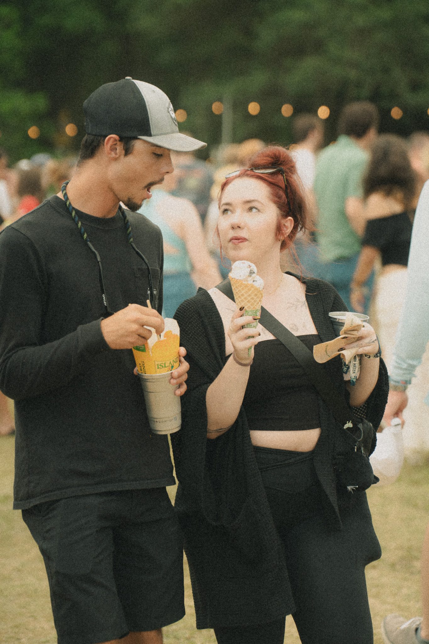 A couple enjoying ice cream at a festival, with a grassy background and other attendees in the distance.