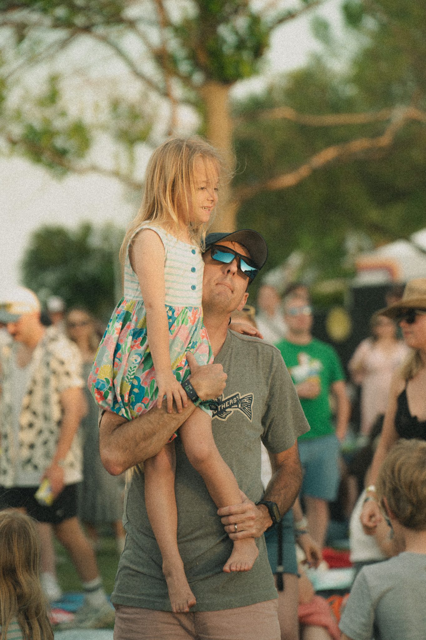 A father holding a young girl on his shoulders at a festival, surrounded by other attendees.