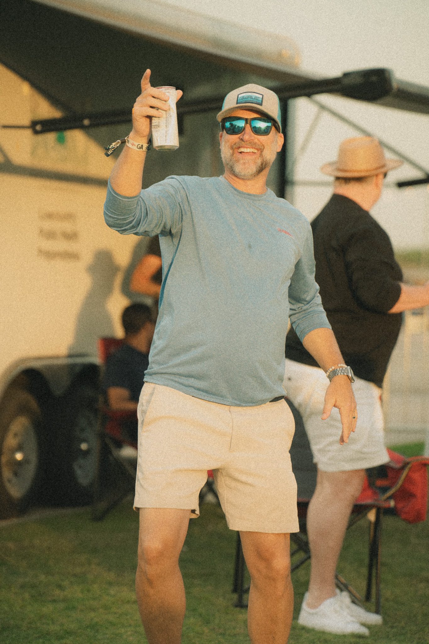 A man holding a can and smiling, wearing sunglasses and a blue long-sleeve shirt, with people and a trailer in the background.