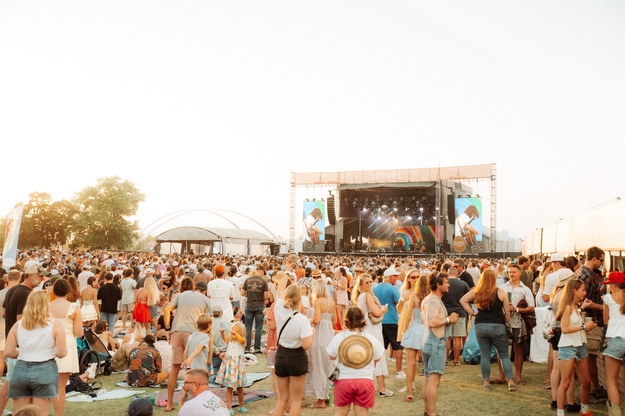 A large crowd enjoying a music festival outdoors, with a prominent stage and performers visible. The sun is setting in the background, creating a warm atmosphere.