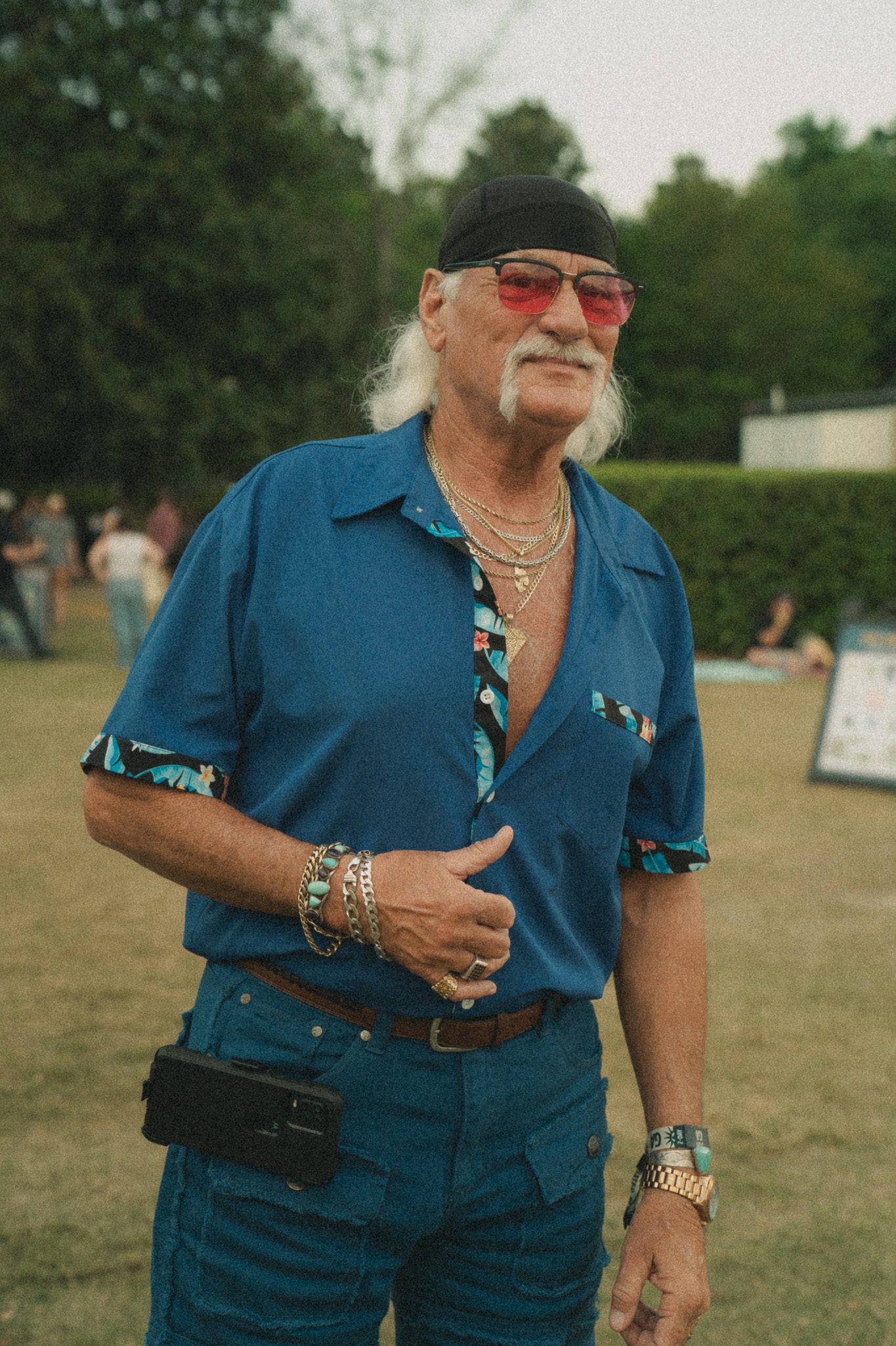 A man with long hair and sunglasses wearing a blue shirt with floral details and background greenery at a festival.