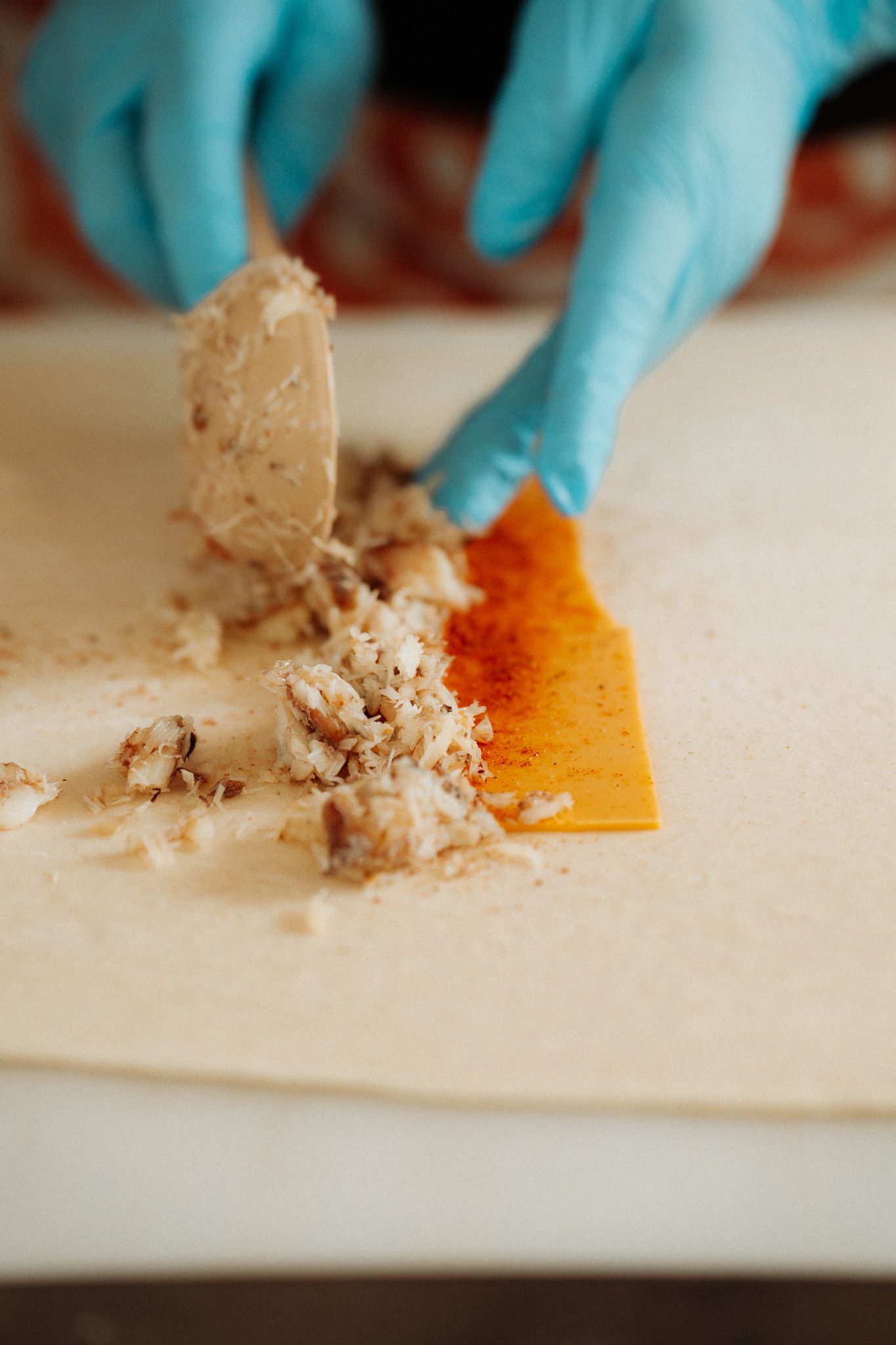 Hands wearing blue gloves preparing a cheese and crab mixture on a flat surface.