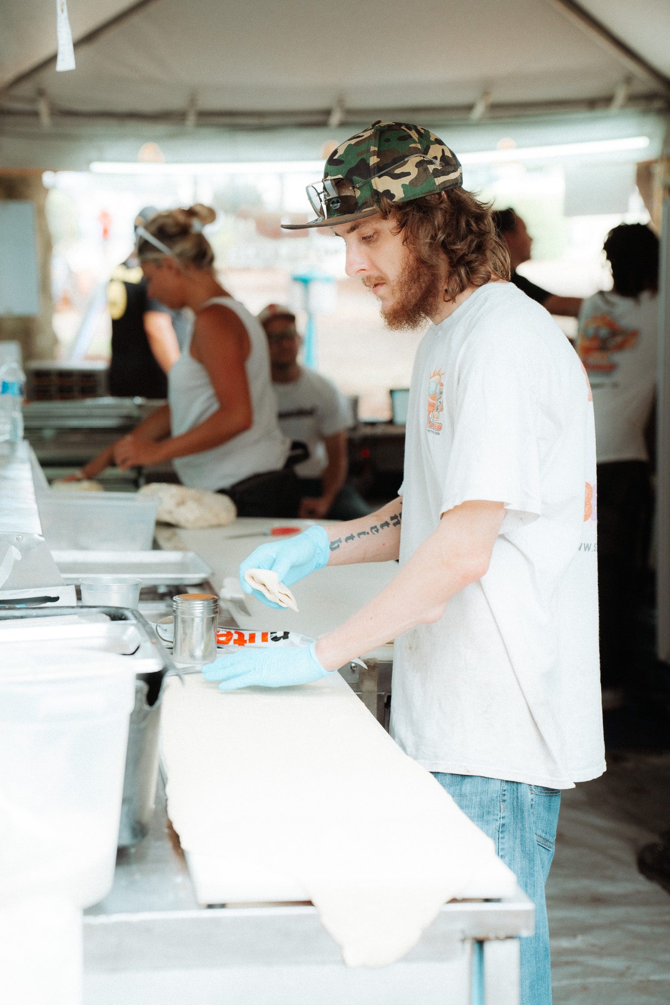 A young man wearing a camouflage hat and gloves is preparing food at a vendor station at a festival, with various food items and other staff members visible in the background.