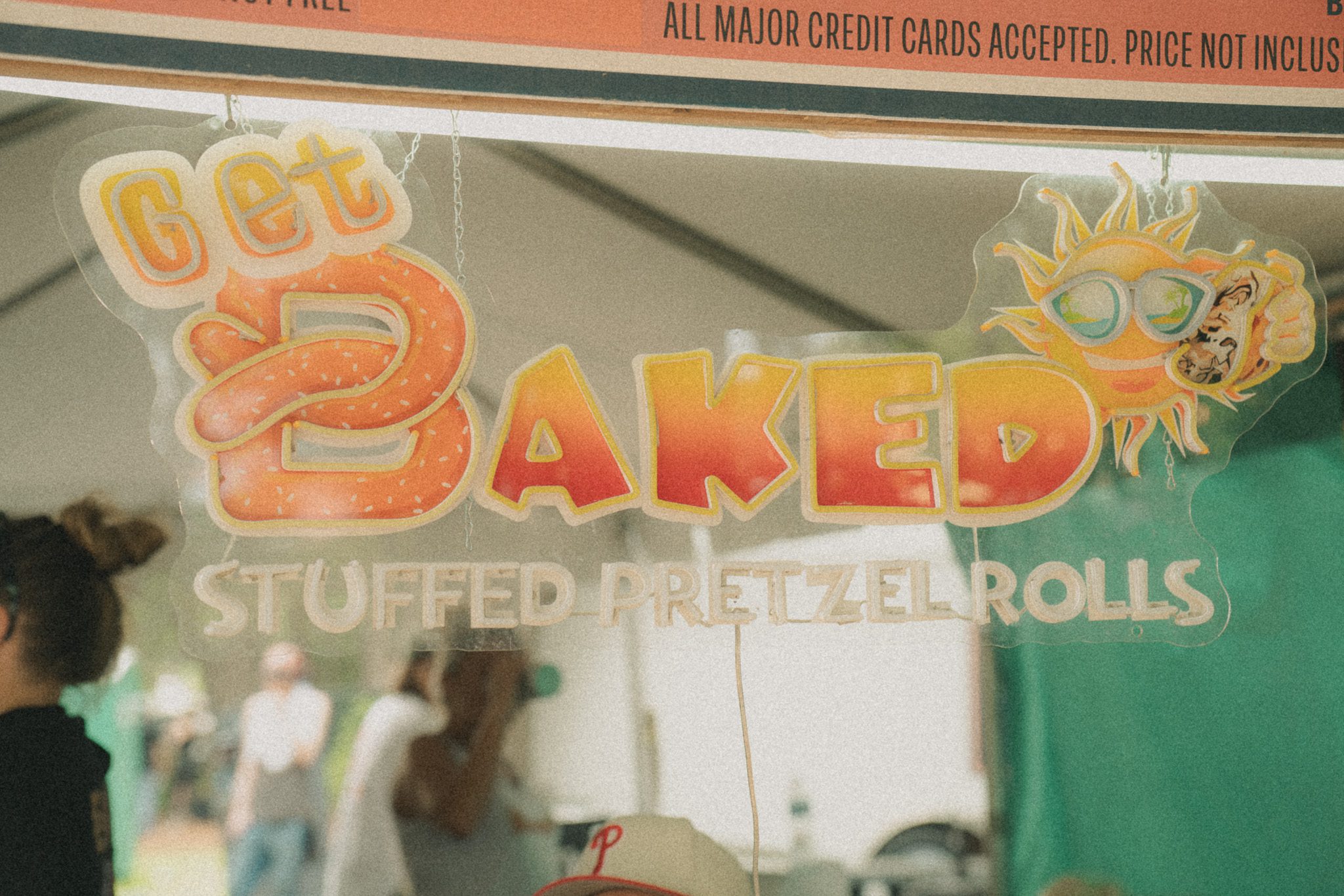 A sign for 'Get Baked' showcasing stuffed pretzel rolls at a festival, with a sun graphic wearing sunglasses.