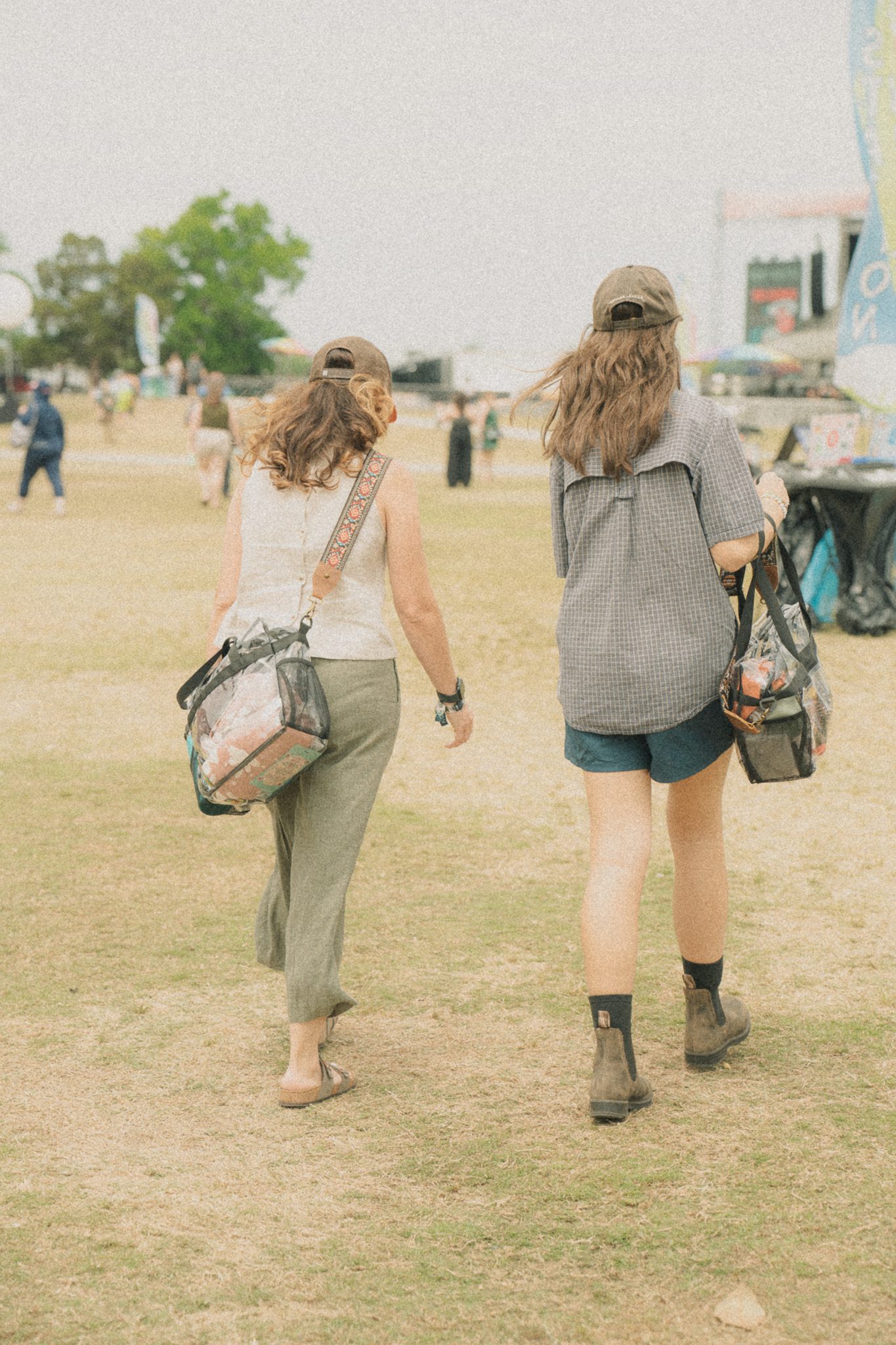 Two women walking side by side on a grassy festival ground, each carrying a bag and wearing caps.