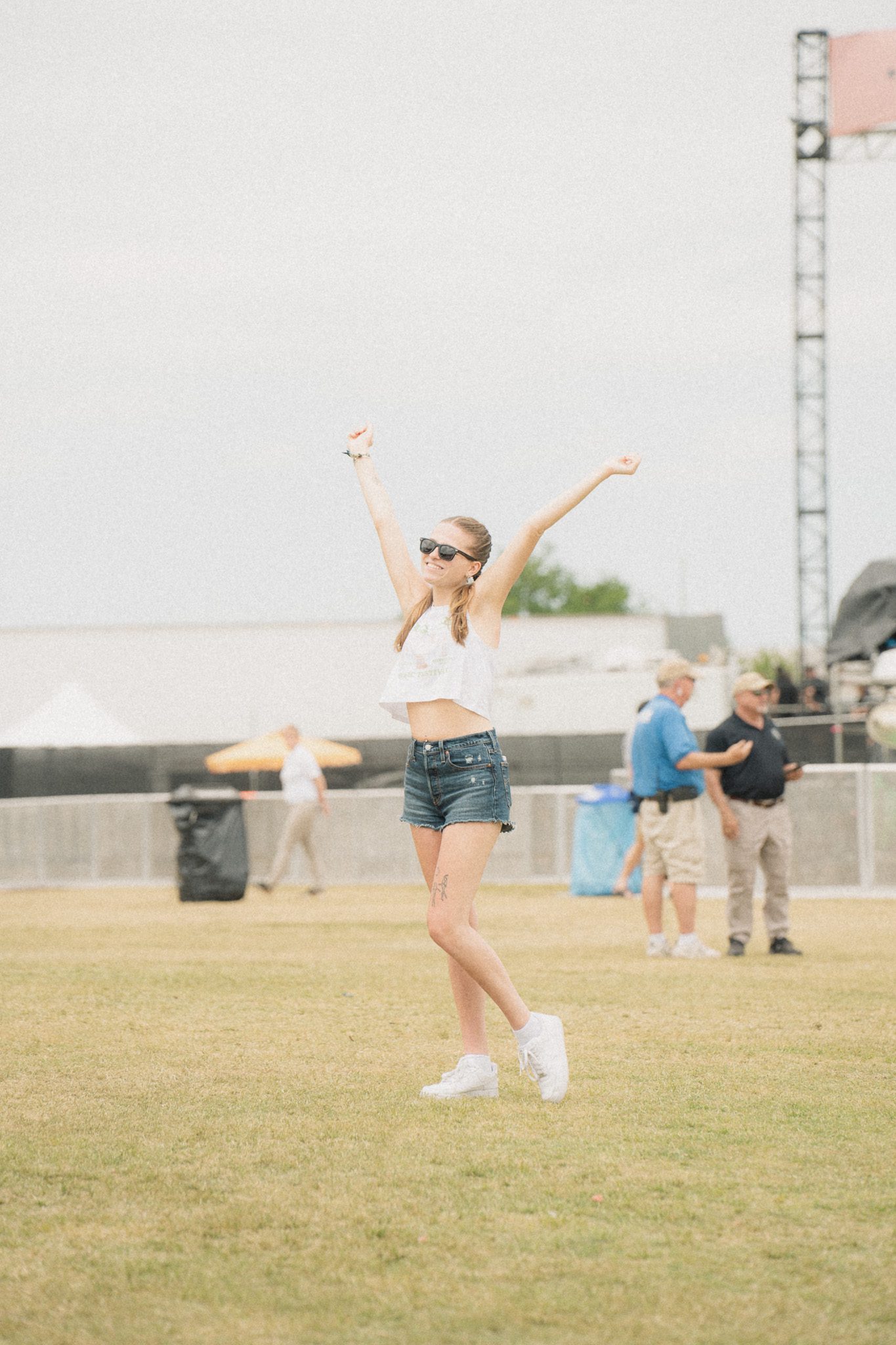 A young woman in a white crop top and denim shorts, smiling and raising her arms in excitement at a festival.