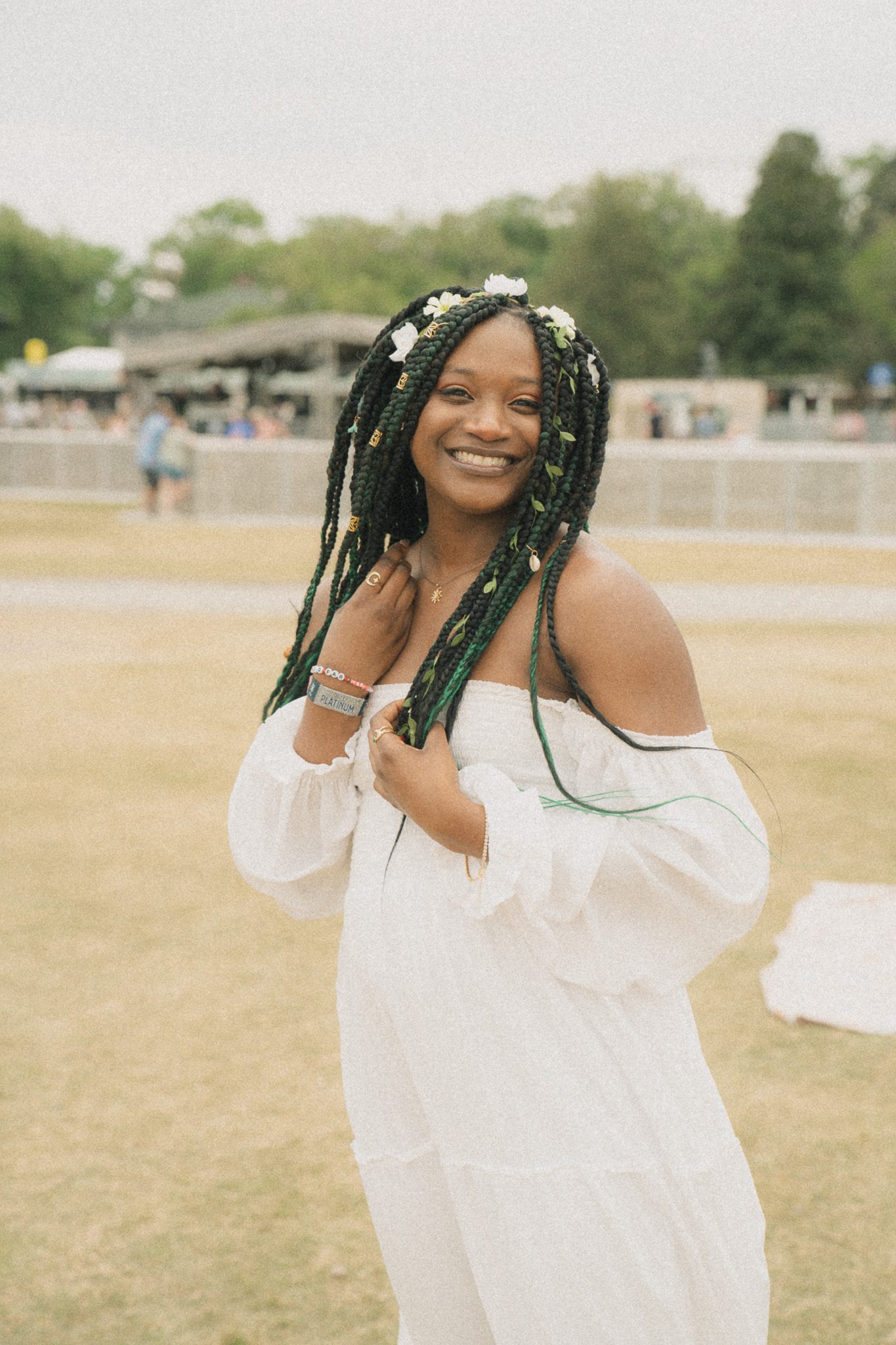 A smiling woman with long braided hair, wearing a white off-shoulder dress, stands outdoors at a festival.