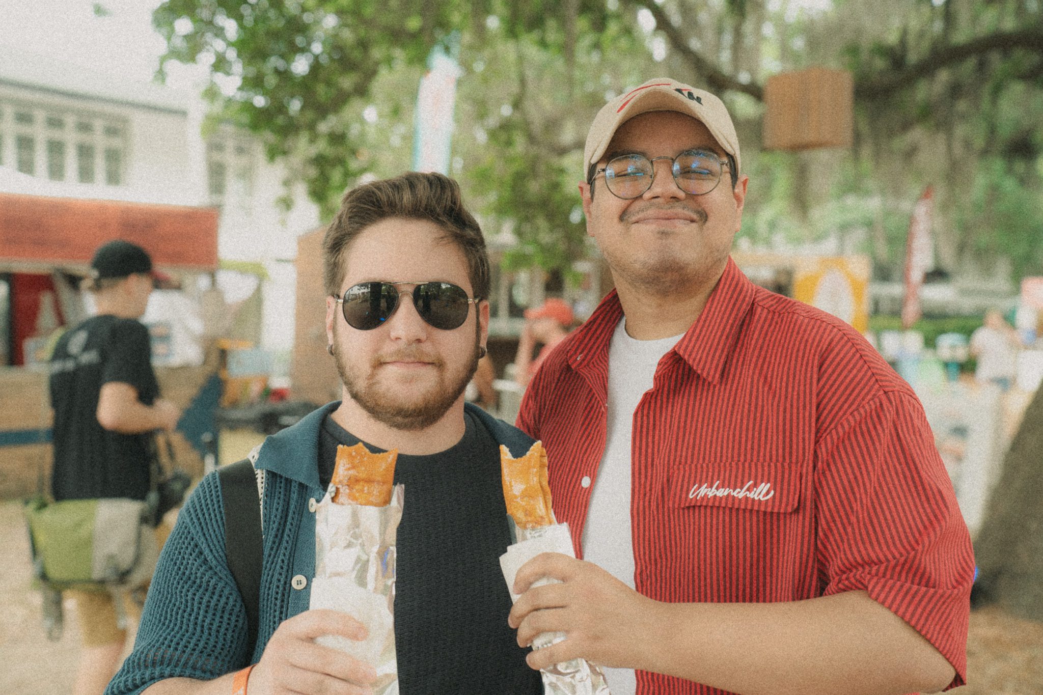Two festival attendees smiling while holding food items at High Water Festival, with festival booths and greenery in the background.
