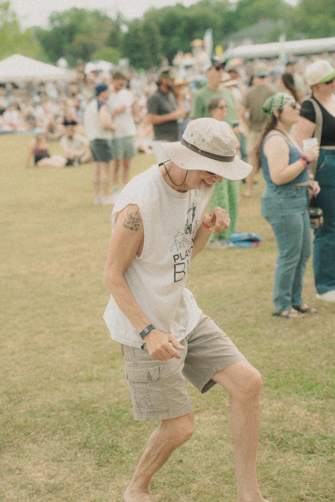 A festival attendee dancing barefoot on grass, wearing a white tank top and a light-colored hat, with a crowd of people in the background.