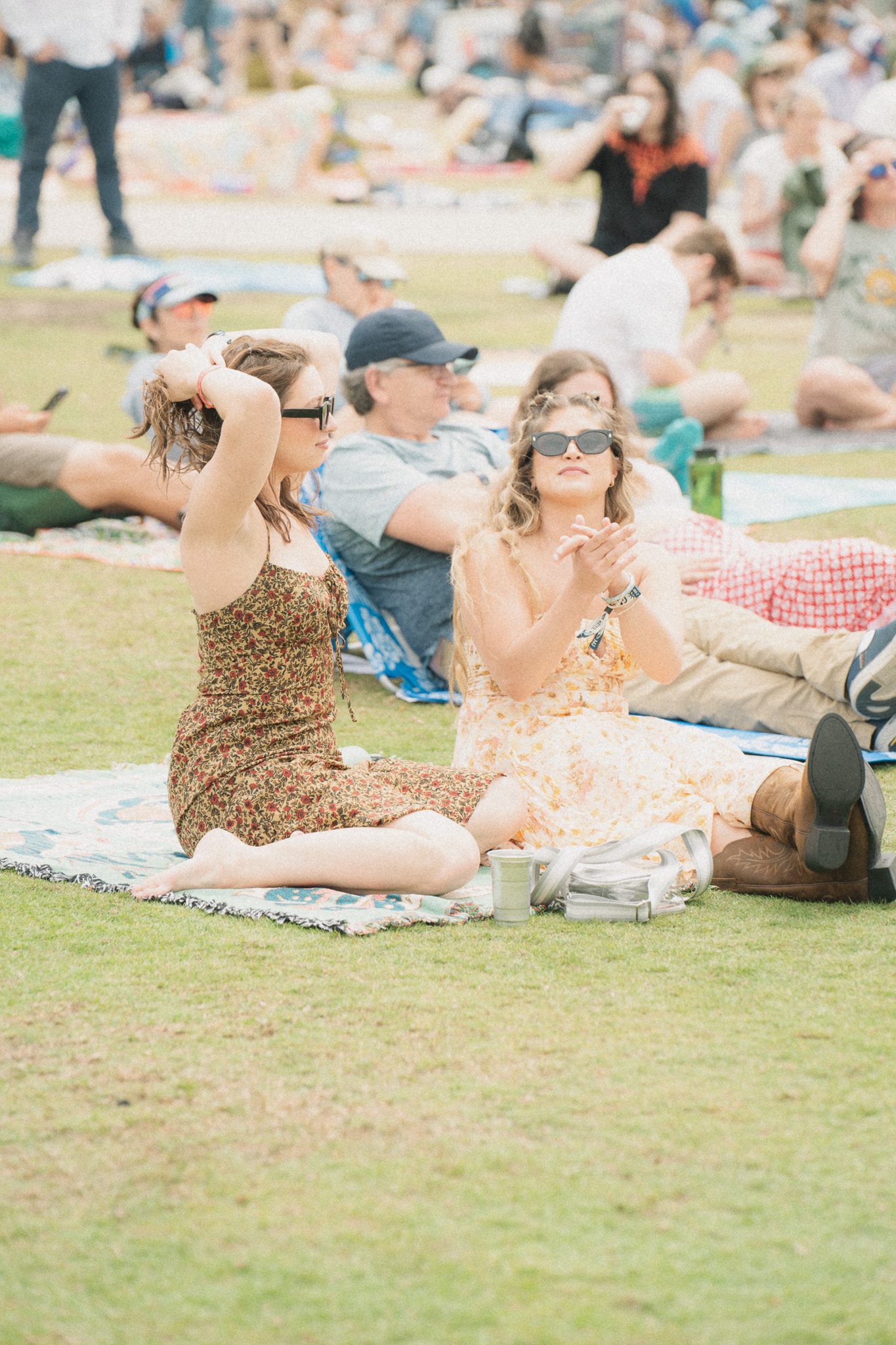 Two women sitting on the grass at a festival, one adjusting her hair and the other clapping, with a crowd of attendees in the background.