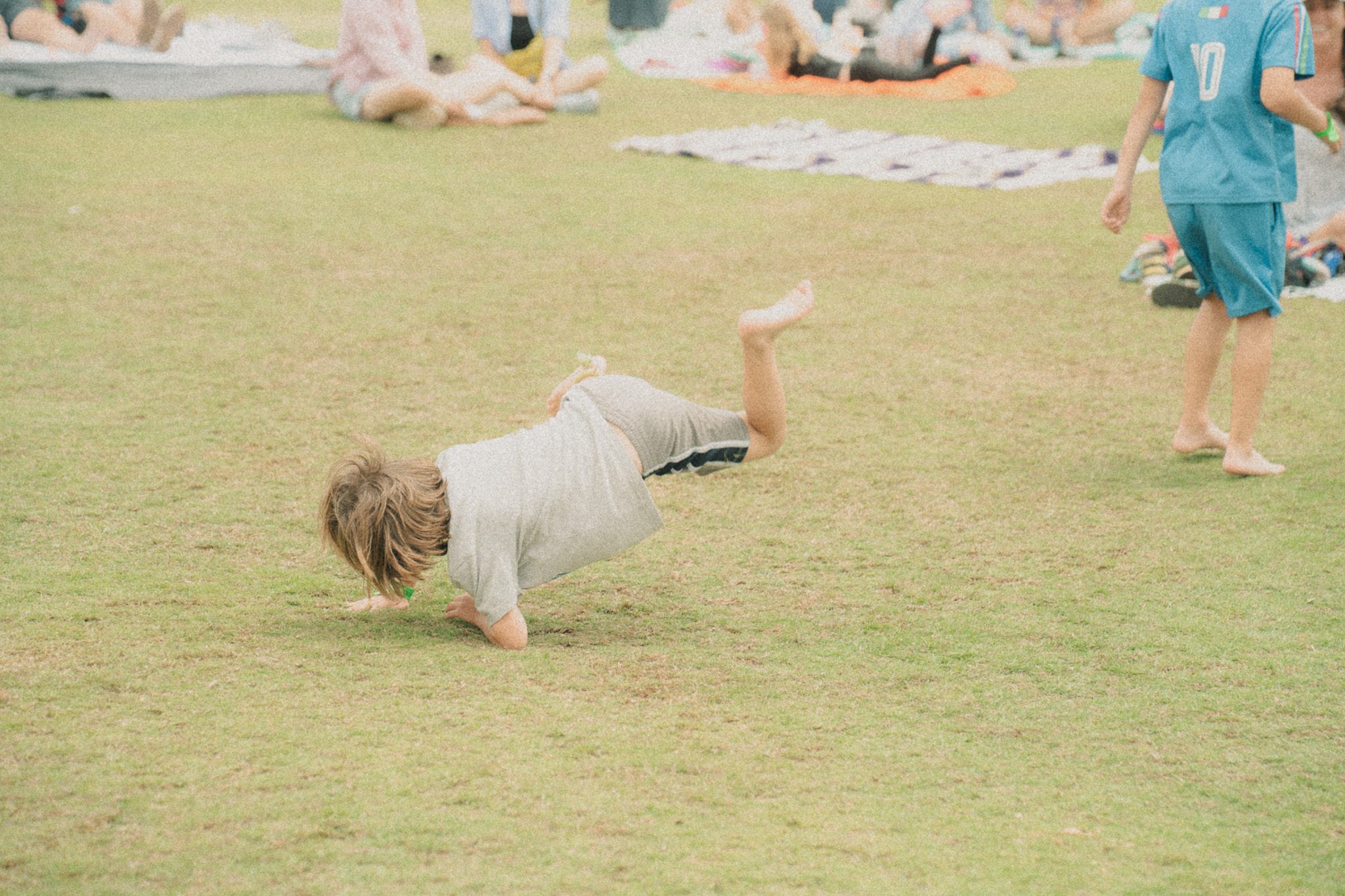 A child playfully rolling on the grass at a festival, surrounded by attendees in the background.