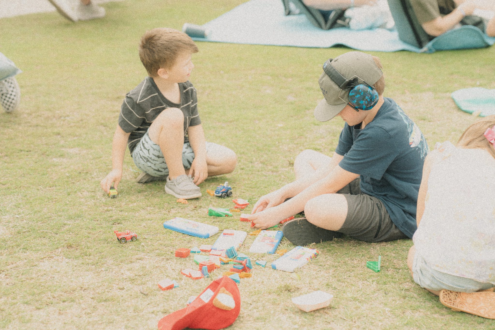 Two young boys playing with toy cars on grass at a festival, one wearing a black striped shirt and the other in a blue shirt with a cap.