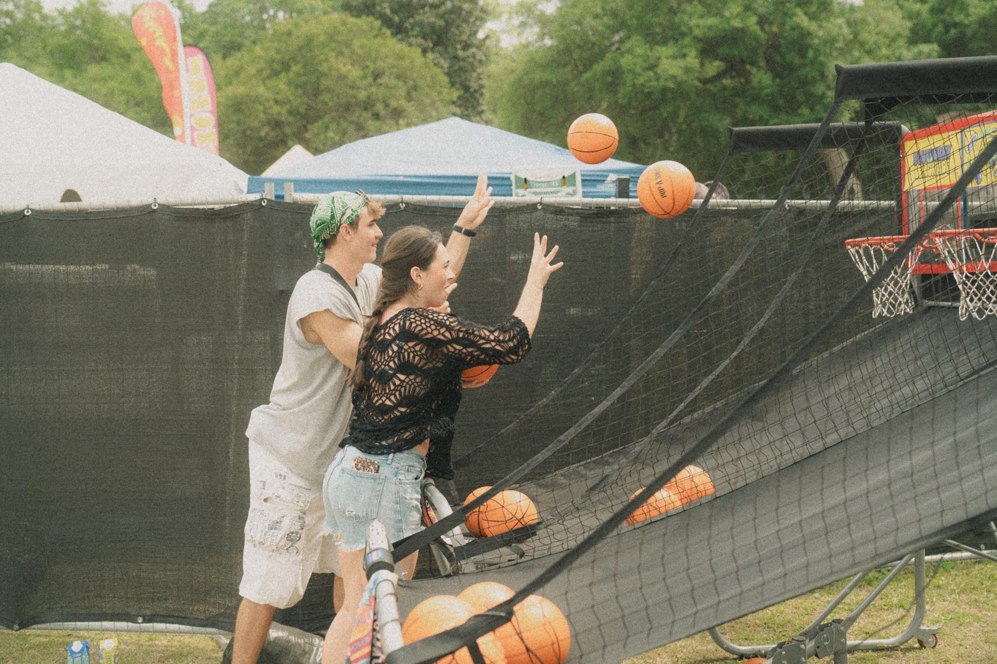 Two festival attendees enjoying a basketball game, throwing basketballs towards a net, surrounded by a vibrant festival atmosphere.