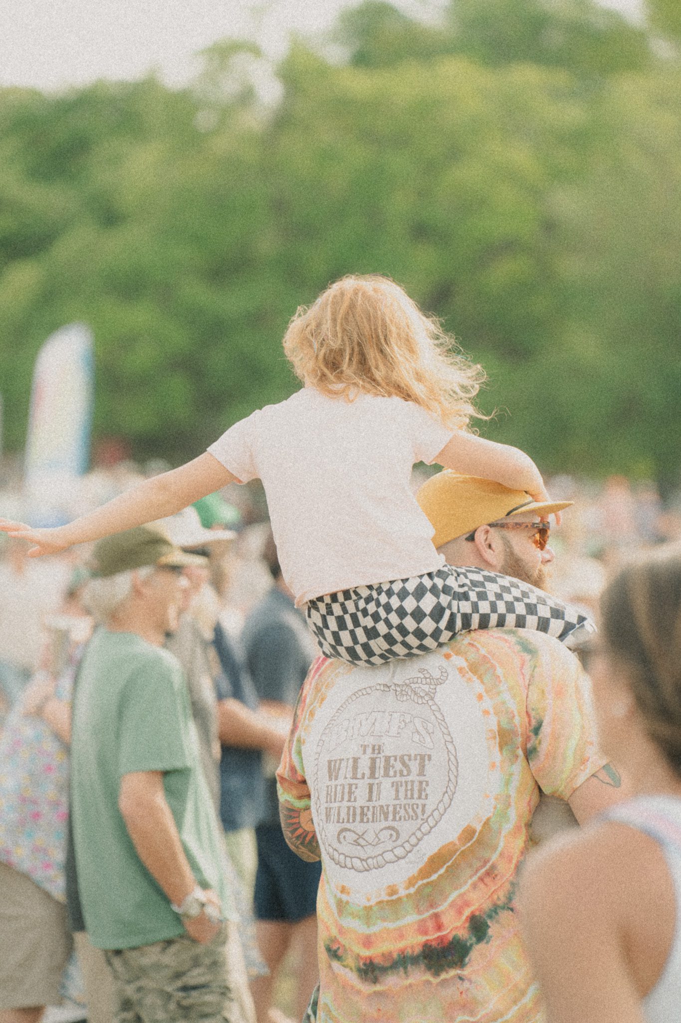 A child with long hair sits on the shoulders of an adult in a colorful shirt, surrounded by festival attendees in a vibrant outdoor setting.