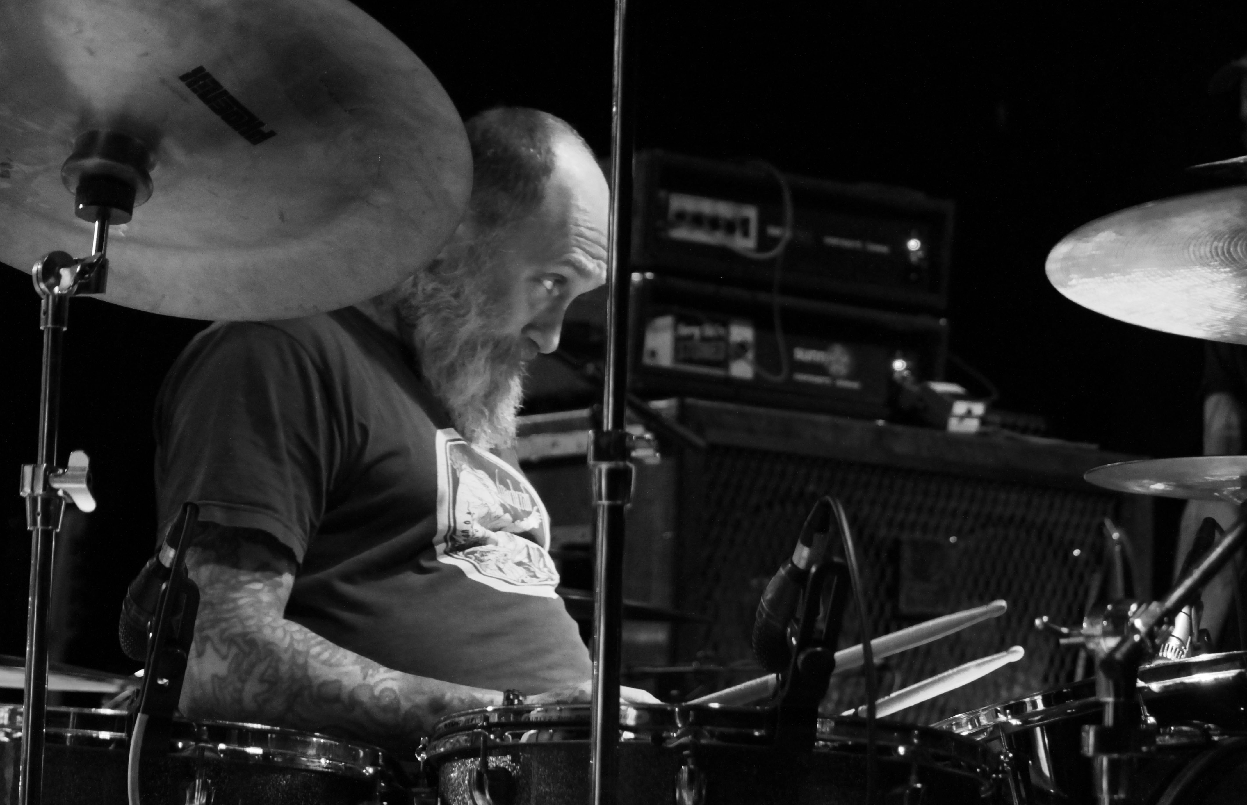 Close-up of a drummer performing on stage, focusing on his intense expression while playing, with a drum kit and amplifiers visible in the background.