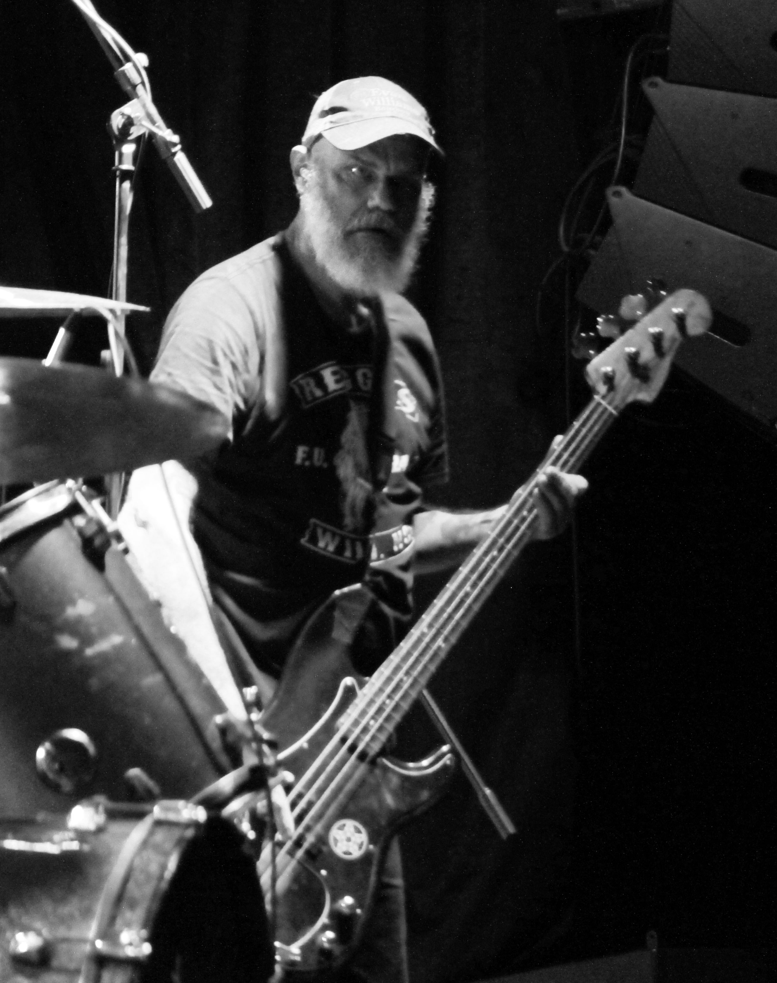 A musician with a beard and a cap playing a bass guitar on stage, with a focused expression, in a black and white setting.