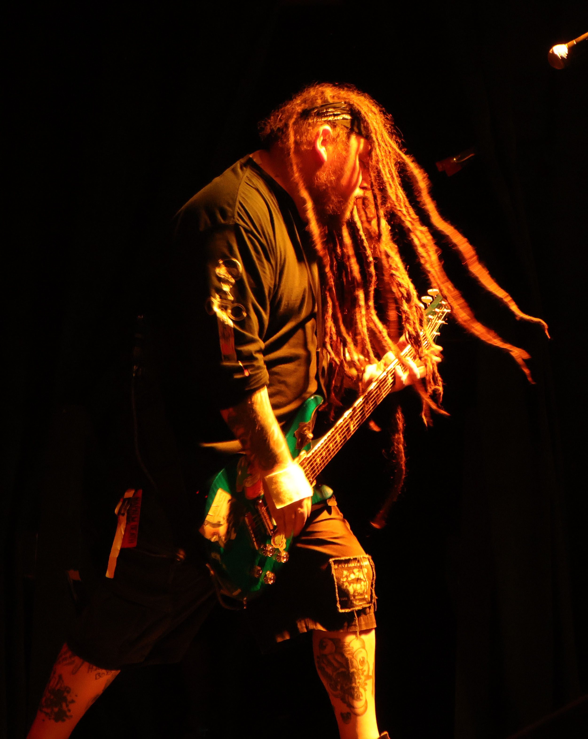 A musician with long dreadlocks performs on stage, energetically playing a green electric guitar under warm lighting.