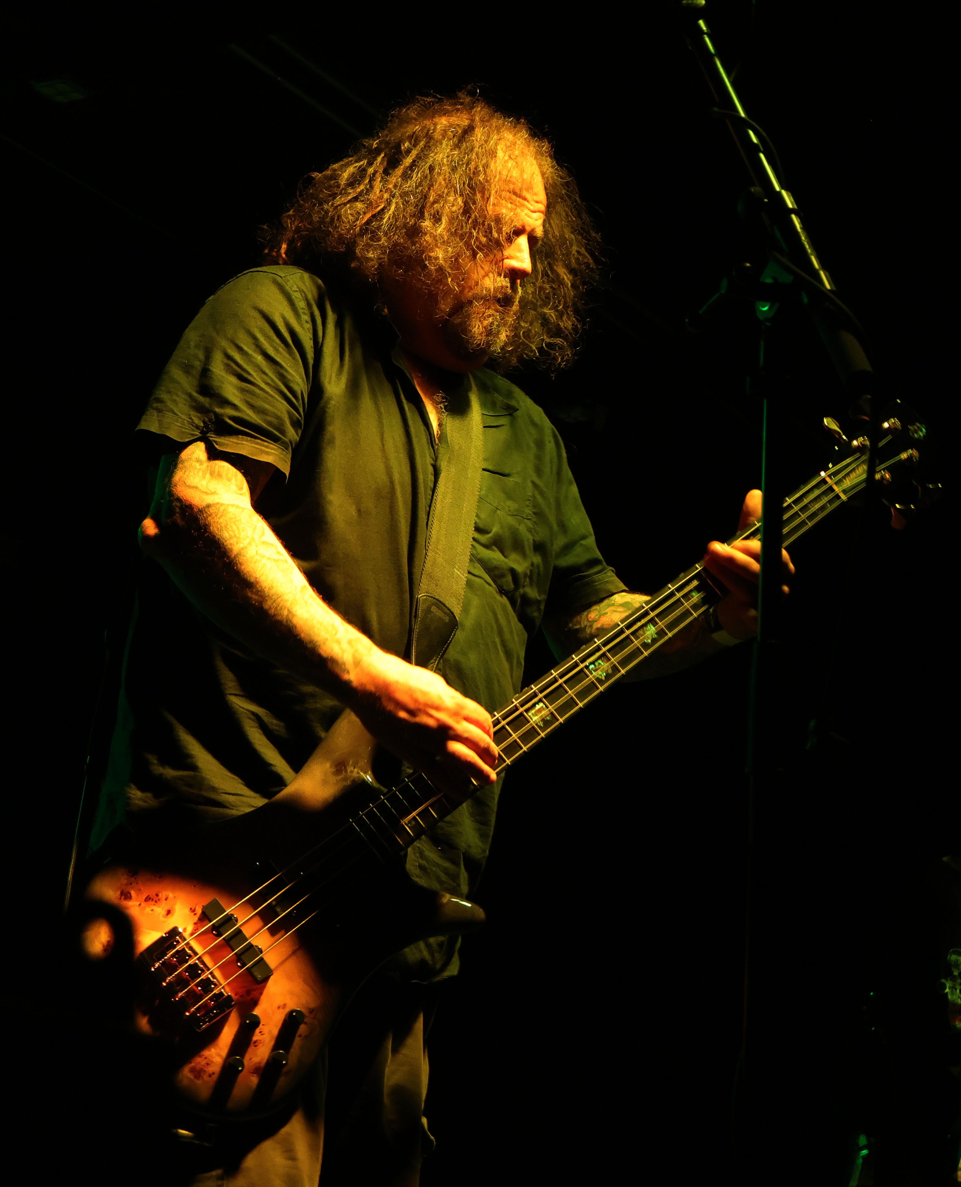 A musician with curly hair passionately playing a bass guitar on stage under green lighting.